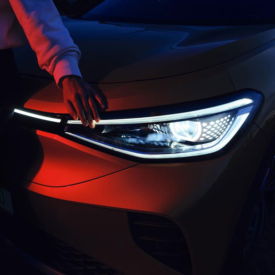 Osoba sa dotýka predného zasvieteného svetlometu v tme. Svetlo automobilu je jasné a detailne prepracované. Fotografiu zdôrazňuje kontrast medzi svetlom a tmavým prostredím.