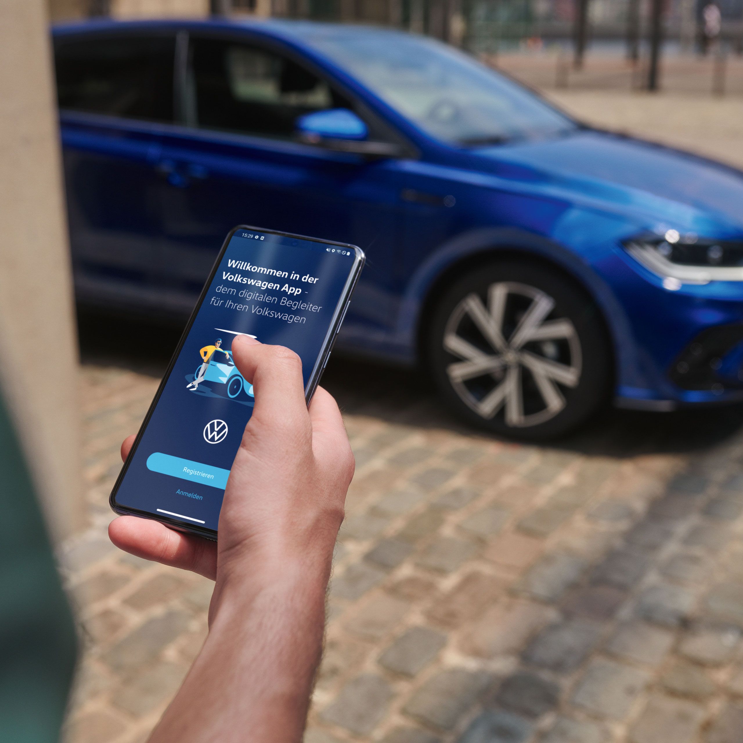 Osoba drží smartfón, na ktorom je otvorená aplikácia Volkswagen. V pozadí vidieť dláždenú cestu, na ktorej je zaparkovaný VW Polo pri budove. 