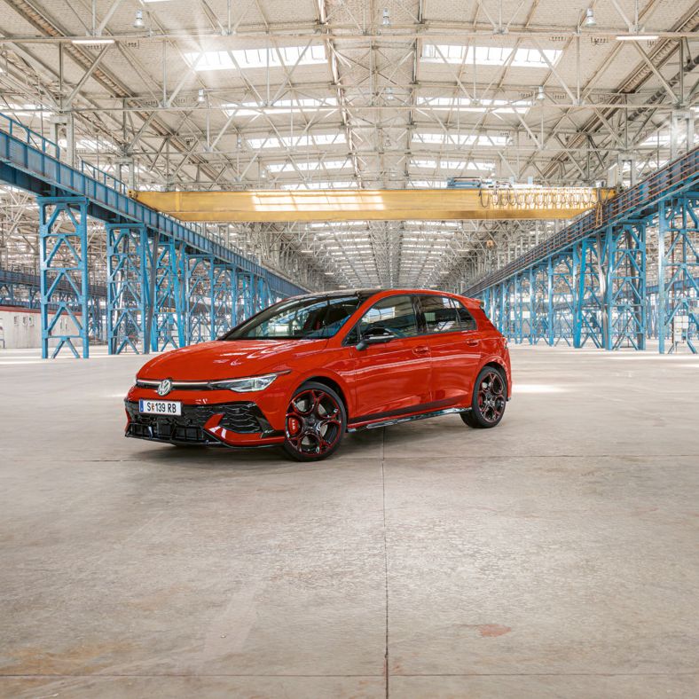 Červený Golf GTI je zaparkovaný vo veľkej, prázdnej priemyselnej budove s modrými oceľovými stĺpmi a nosníkmi. Strecha budovy je zo skla a prepúšťa dovnútra dostatok svetla.