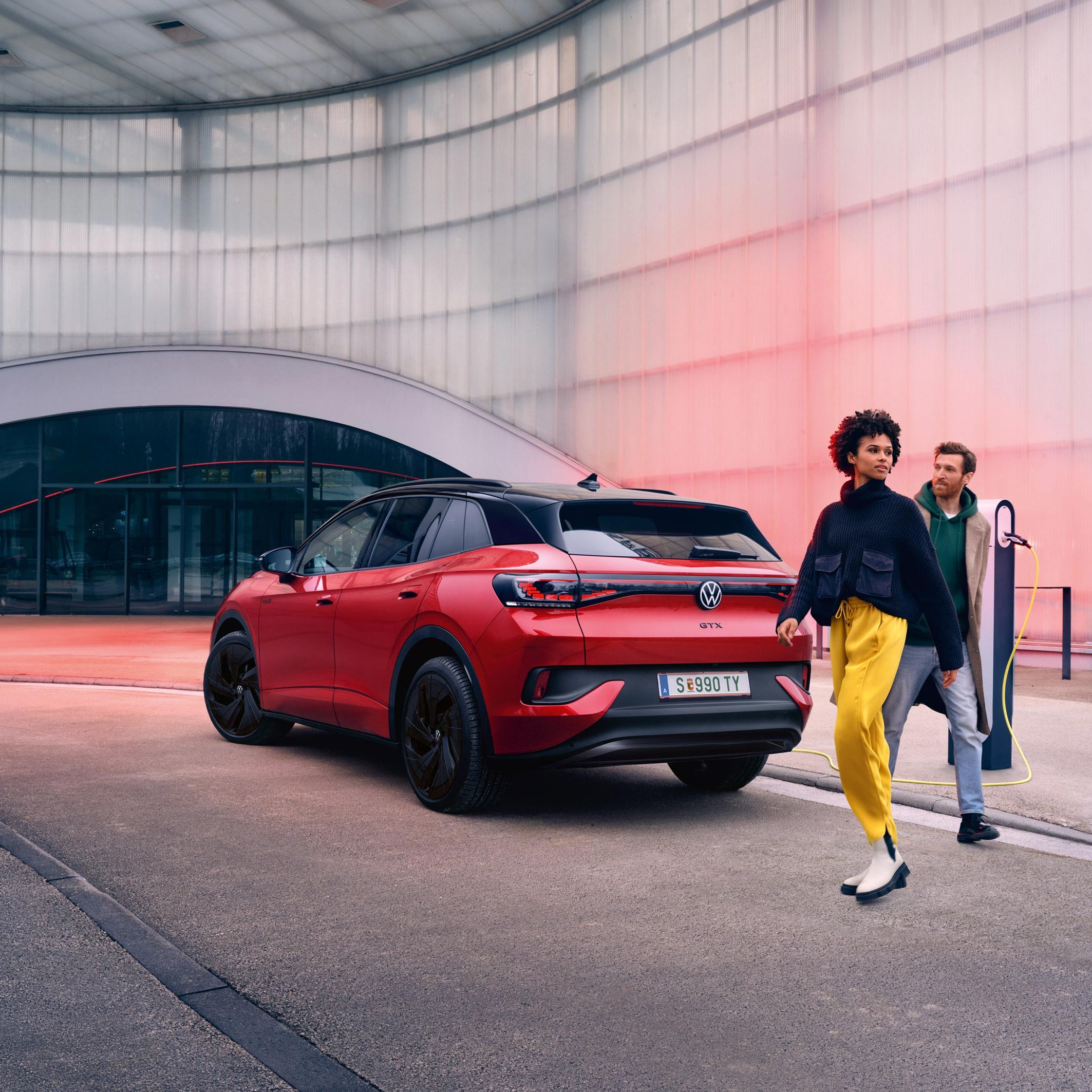 Pohľad na červený VW ID.4 GTX zozadu a zboku. Nabíja sa pri ceste, žena a muž prechádzajú okolo. Stojí pri  modernej budove s priehľadnou fasádou.