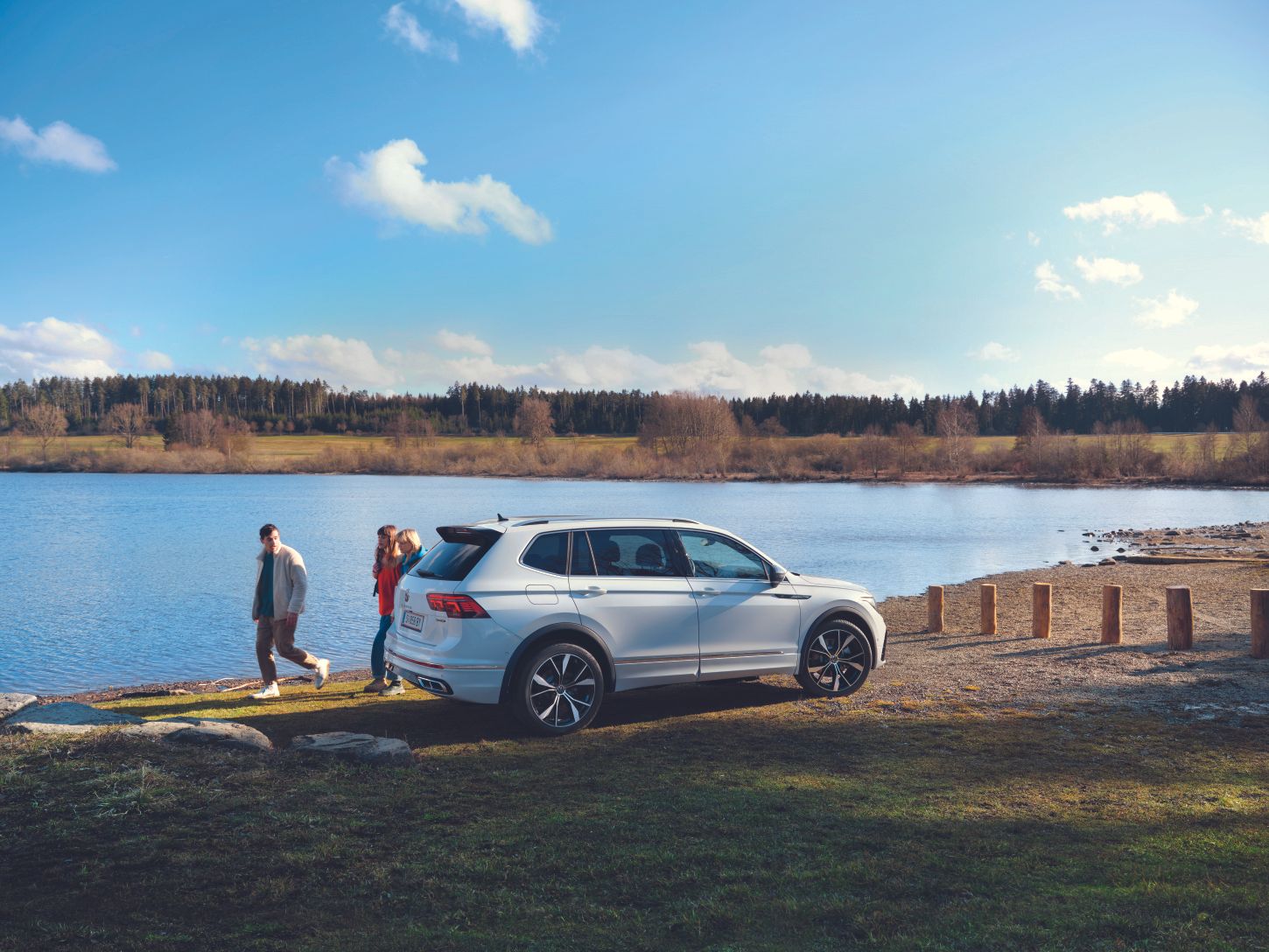 VW Tiguan Allspace stojí pri jazere, za ním prechádzajú traja ľudia. Jazero obklopuje zeleň a les.