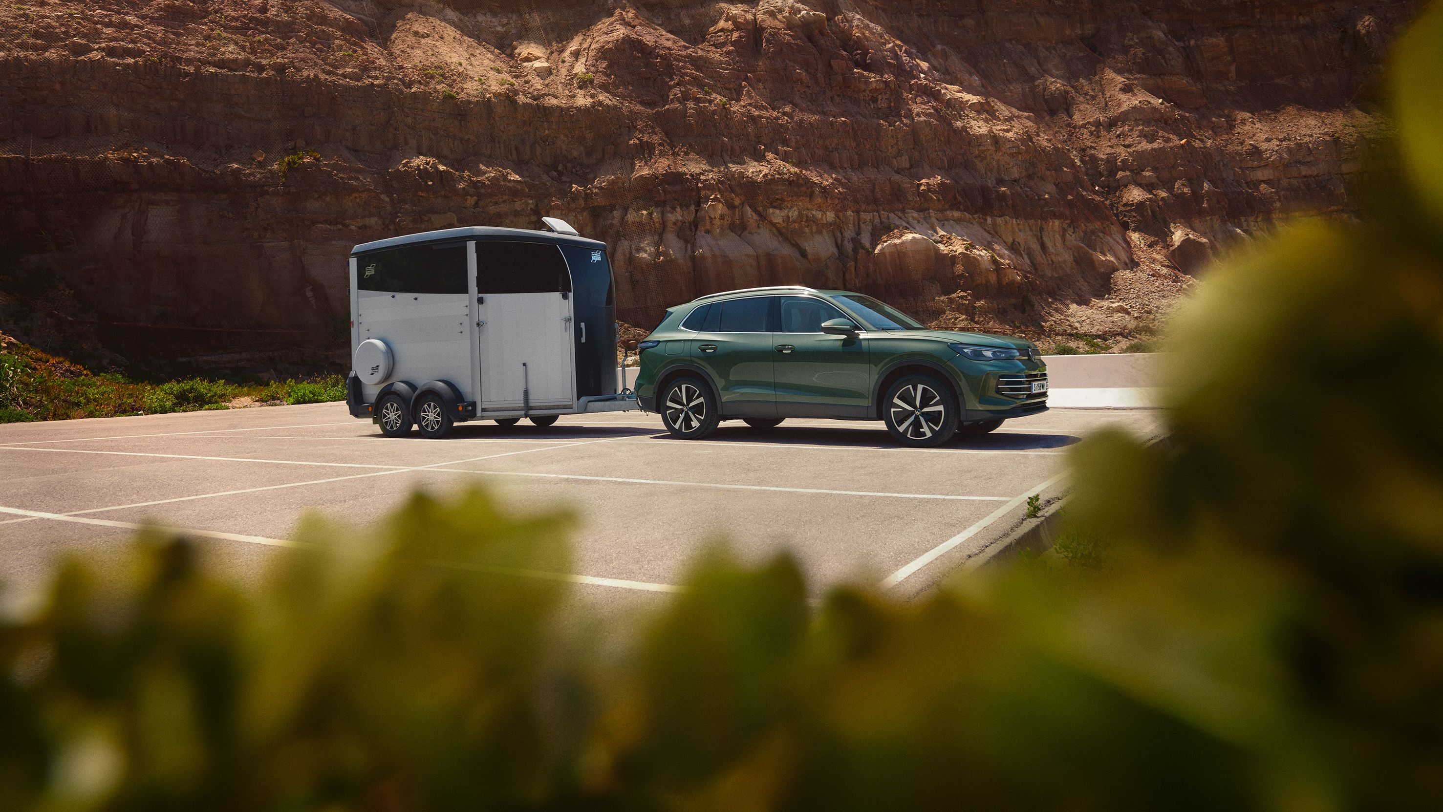 Zelené SUV auto je zaparkované na parkovisku s pripojeným prrívesom. Pozadie tvorí vysoký kamenný útes. V popredí sú rozostrené zelené listy stromov.