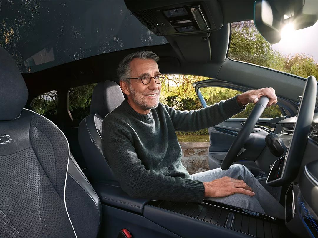 Muž sedí v modernom aute, drží volant a má otvorené dvere. Interiér auta je elegantný s pohodlnými sedadlami. Von vidieť prírodu.