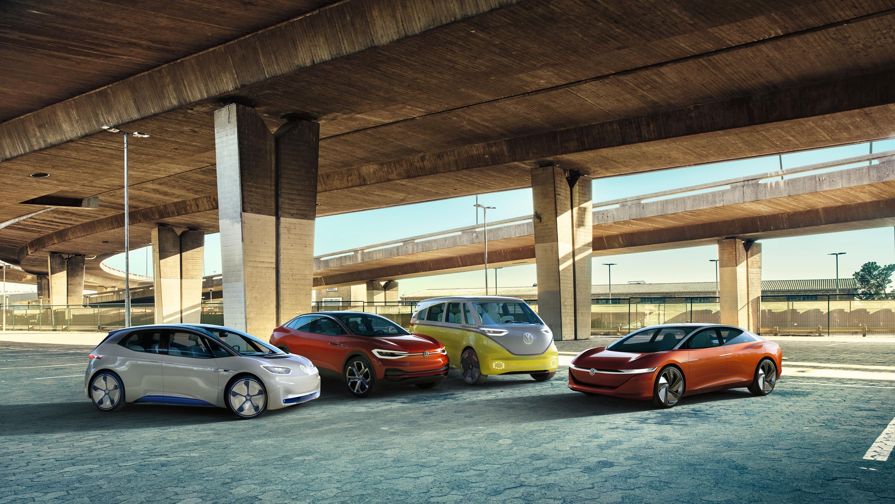 Pod mostom je zaparkovaných niekoľko moderných elektrických vozidiel Volkswagen rôznych farieb. Miesto pôsobí prázdne a má priemyselný nádych.