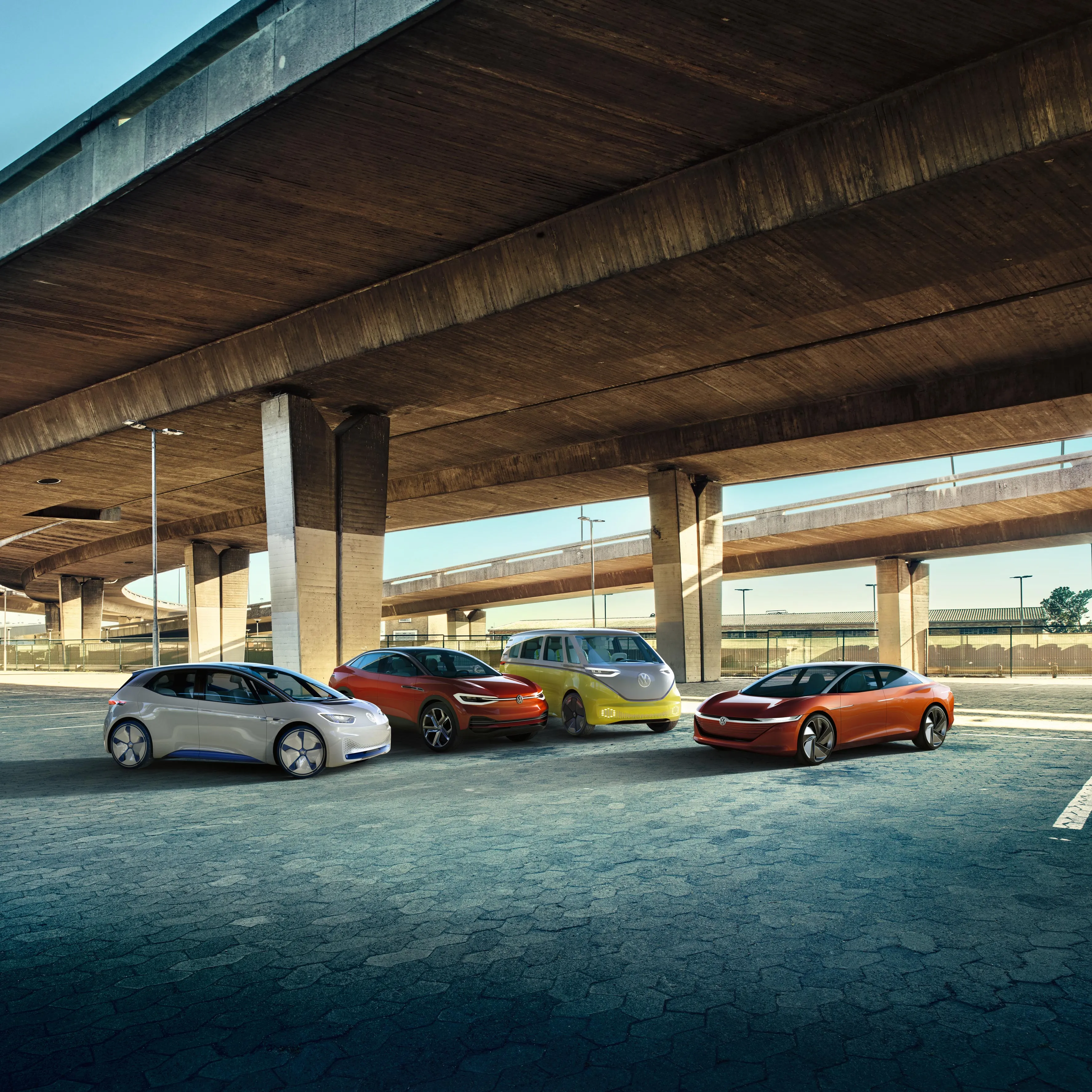 Pod mostom je zaparkovaných niekoľko moderných elektrických vozidiel Volkswagen rôznych farieb. Miesto pôsobí prázdne a má priemyselný nádych.
