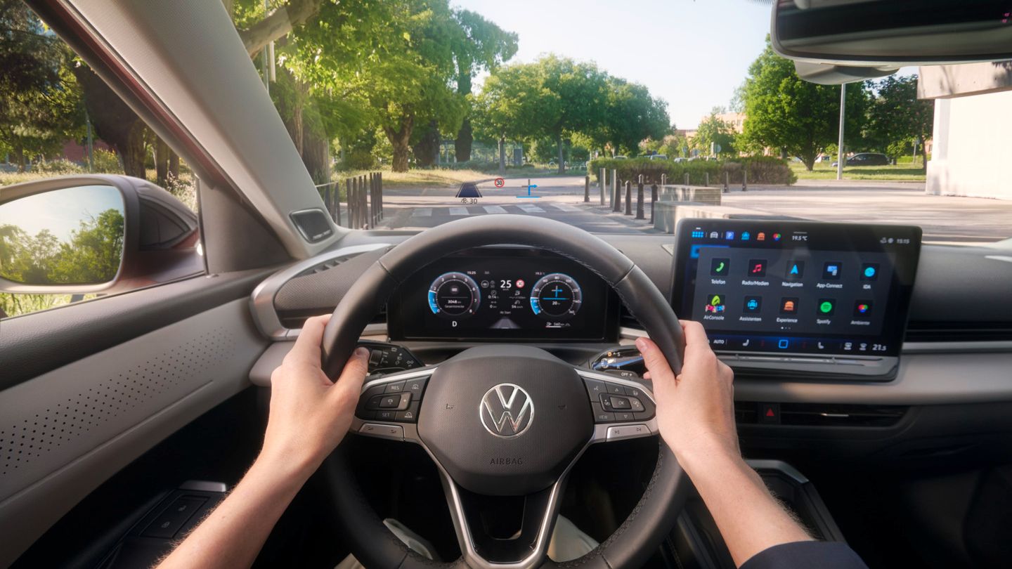 Pohľad na prístrojový panel a displej MMI v VW T-Roc Style s detailom na head-up displej na čelnom skle. Cez čelné sklo vidieť cestu, vpravo modernú, verejnú budovu a vľavo mestský park..