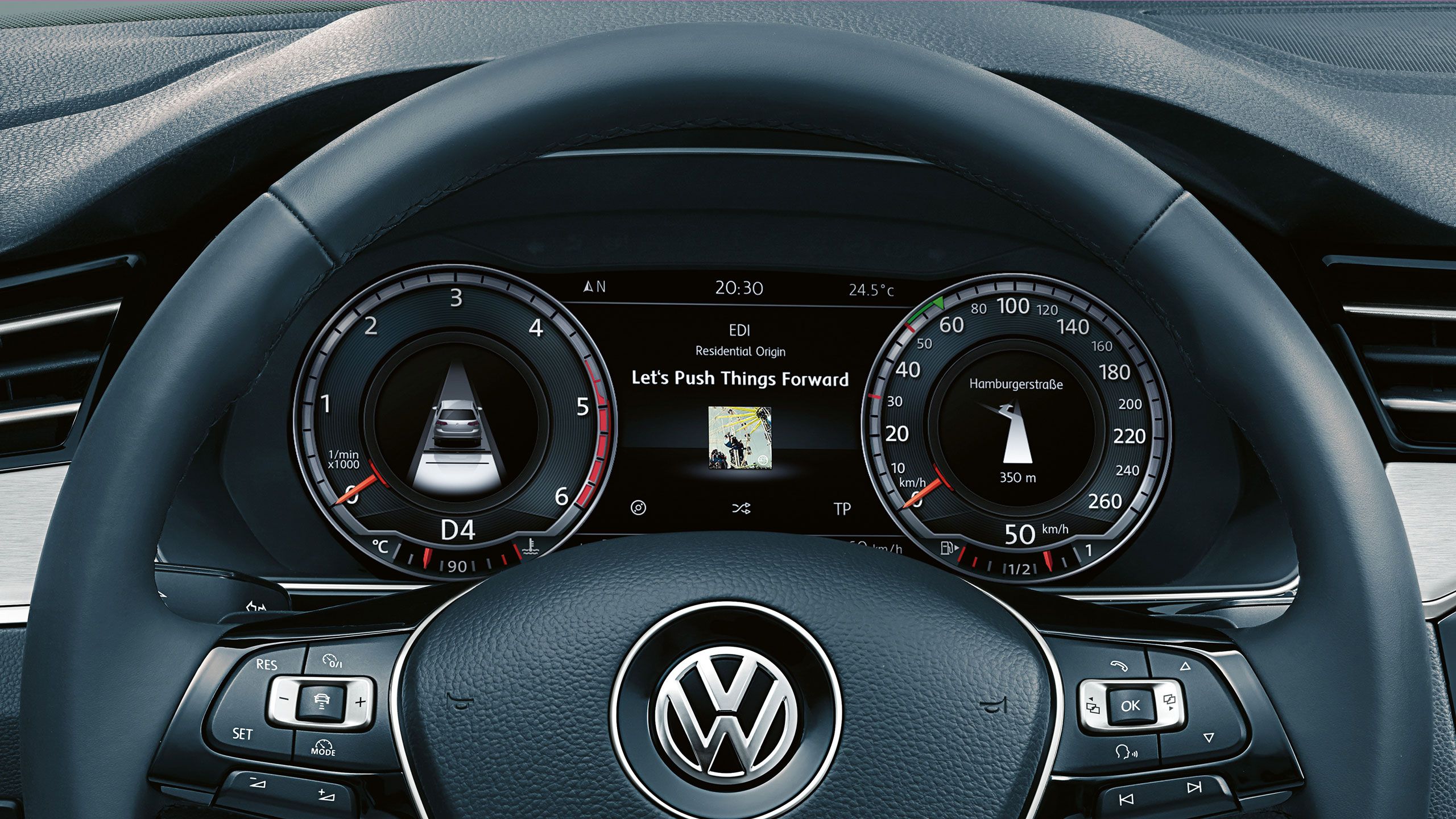 Volant automobilu značky Volkswagen, za ním moderný digitálny prístrojový panel. Na displeji je informácia o prehrávanej piesni. Na volante je vidieť tlačidlá na ovládanie funkcií.