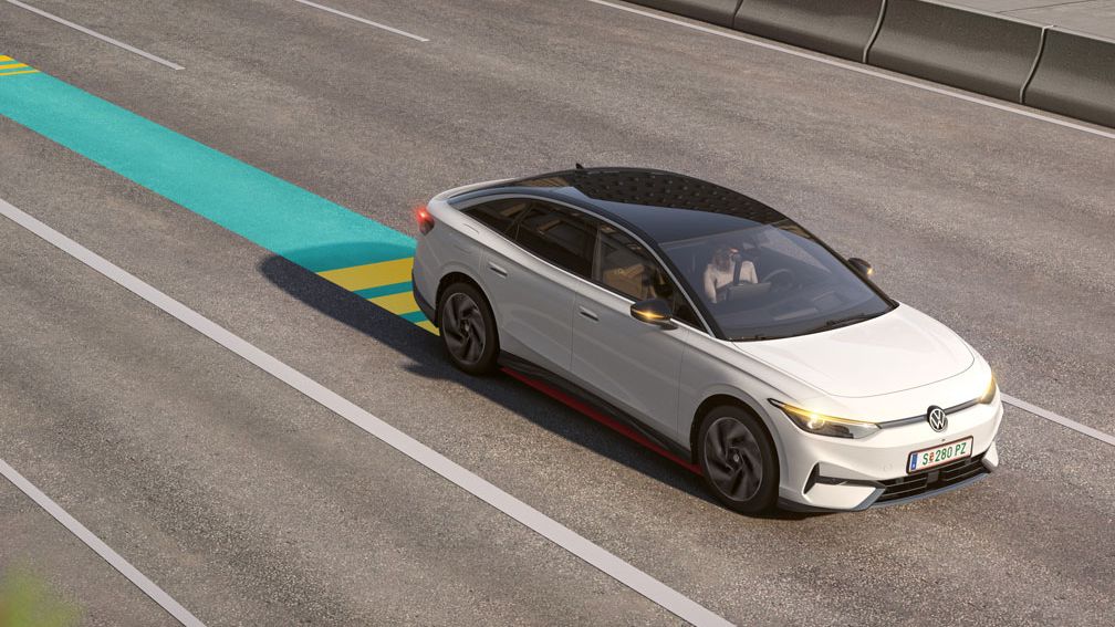 Vozidlo jazdiace po diaľnici, nasledované ďalšími autami. Na ceste je vyznačený modro-žltý pruh, ktorý graficky vysvetľuje systém Emergency Assist.