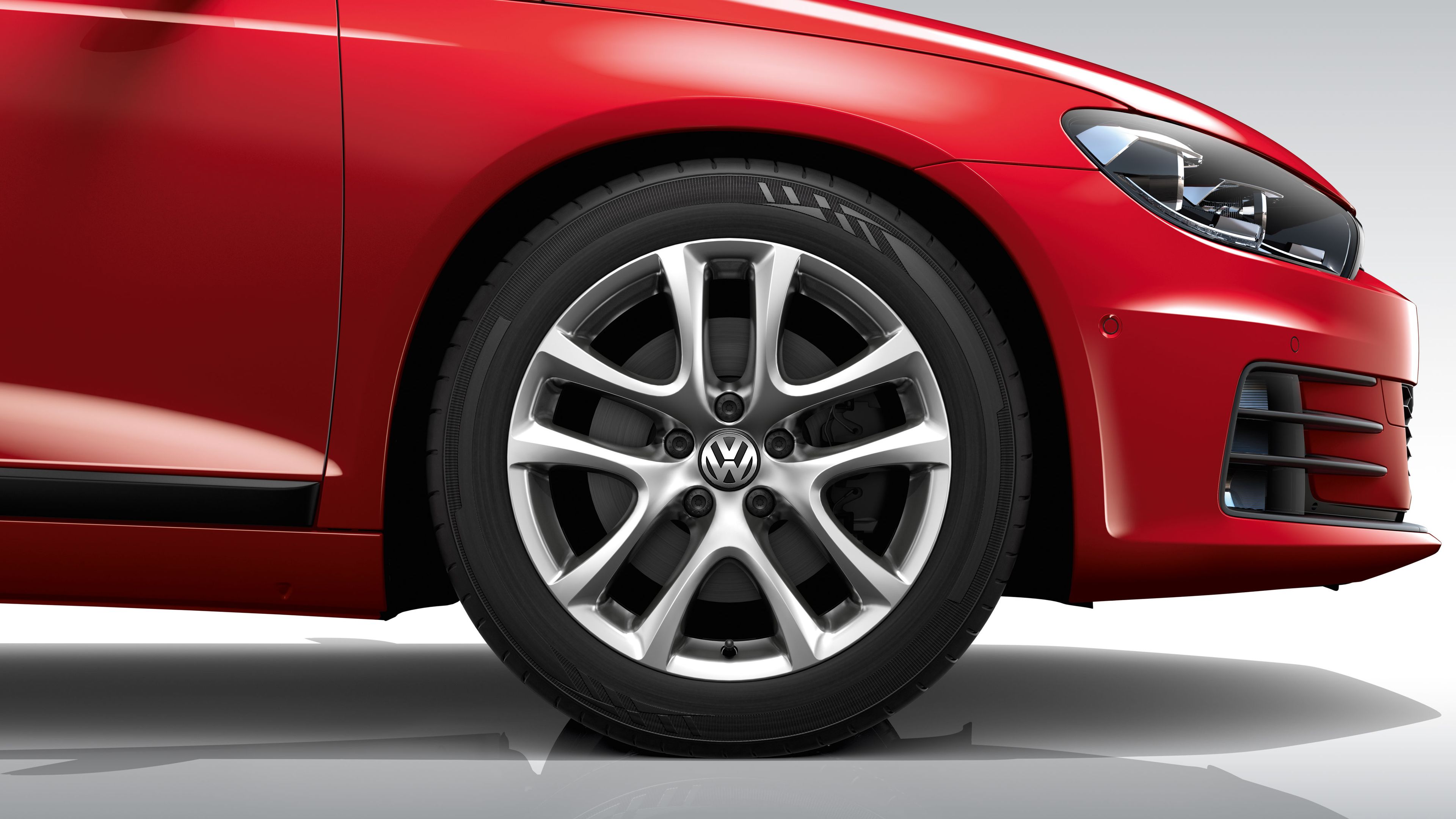 Detailný pohľad na koleso červeného automobilu Volkswagen. Viditeľný je predný blatník a predná časť vozidla. Koleso má disk z ľahkej zliatiny s piatimi lúčmi.