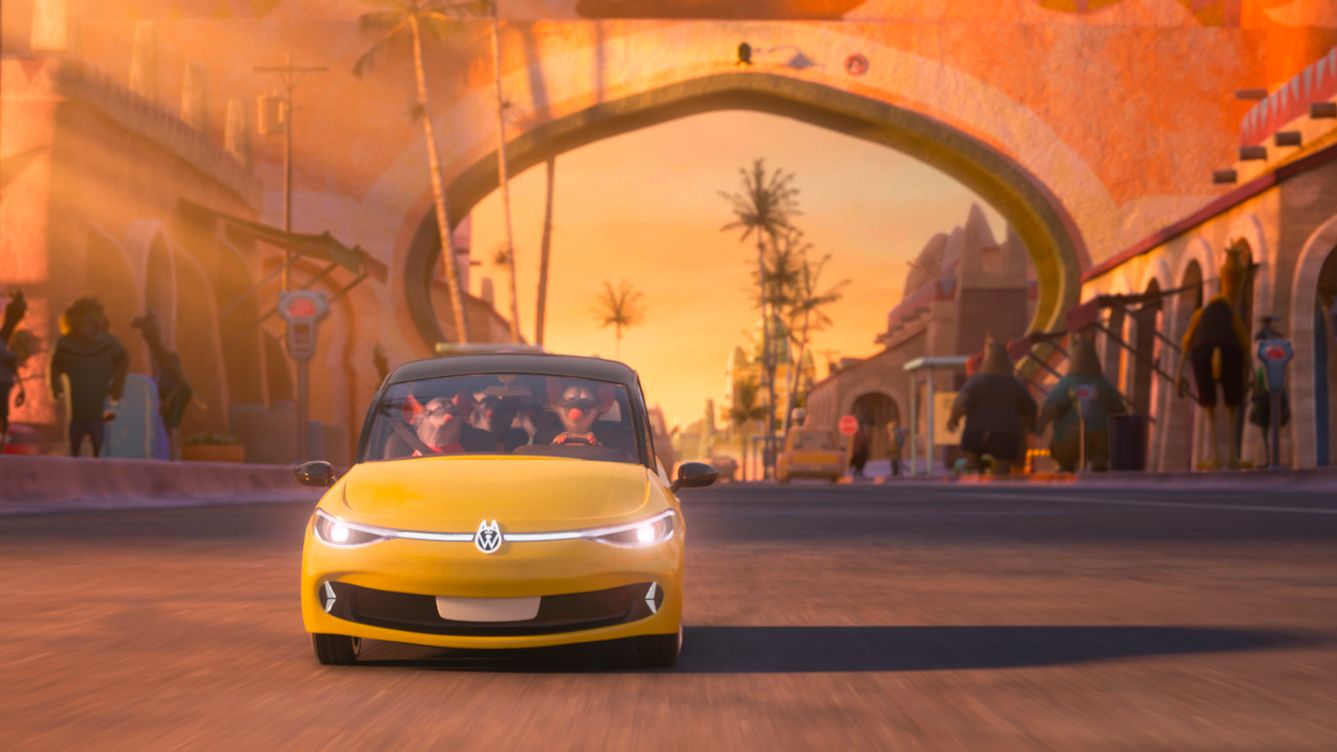 Animované auto žltej farby jazdí po historickej ulici v exotickom meste. Vo vozidle sú dvaja pasažieri. Na ulici sa nachádzajú ďalší ľudia a stánky.