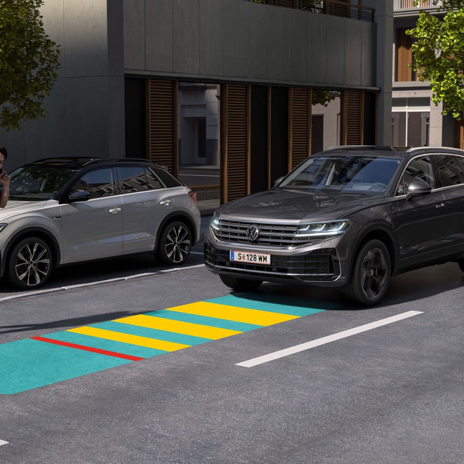 VW Volkswagen Touareg jazdí po ceste, chlapec prechádza cez cestu a pozerá sa na opačnú stranu, takže nevníma približujúce sa vozidlo. Modro-žlto-červenou šípkou je graficky znázornený adaptívny tempomat. Scéna sa odohráva v meste. 