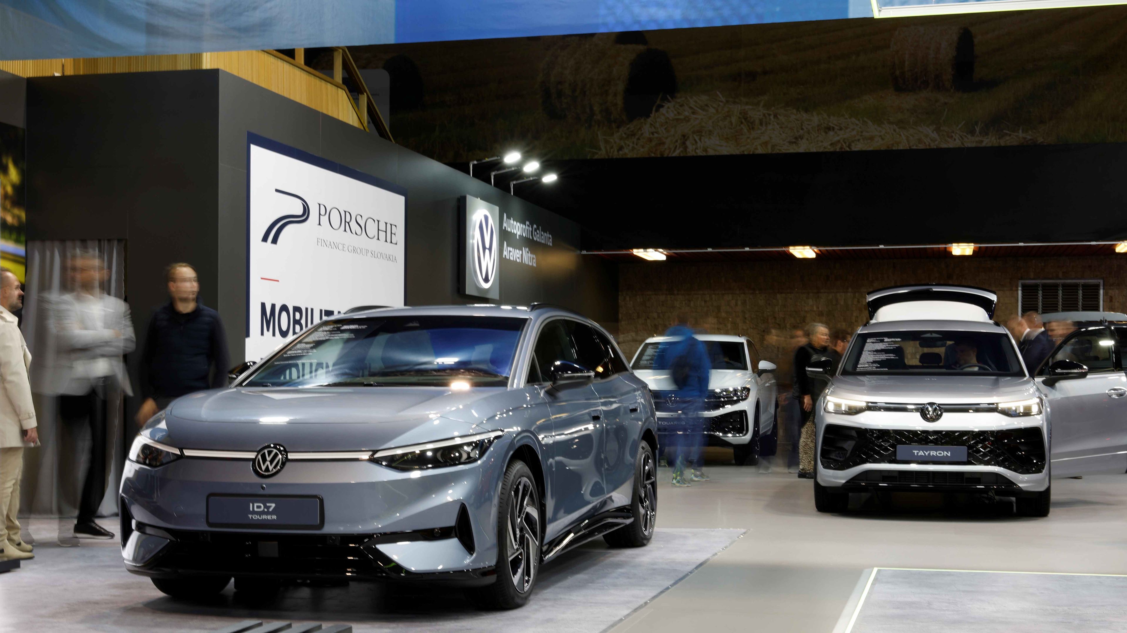Na výstave sú prezentované elektrické vozidlá značiek Volkswagen. V popredí je modrý Volkswagen ID.7 a v pozadí biele SUV s otvoreným batožinovým priestorom. Okolo áut sa pohybujú návštevníci.