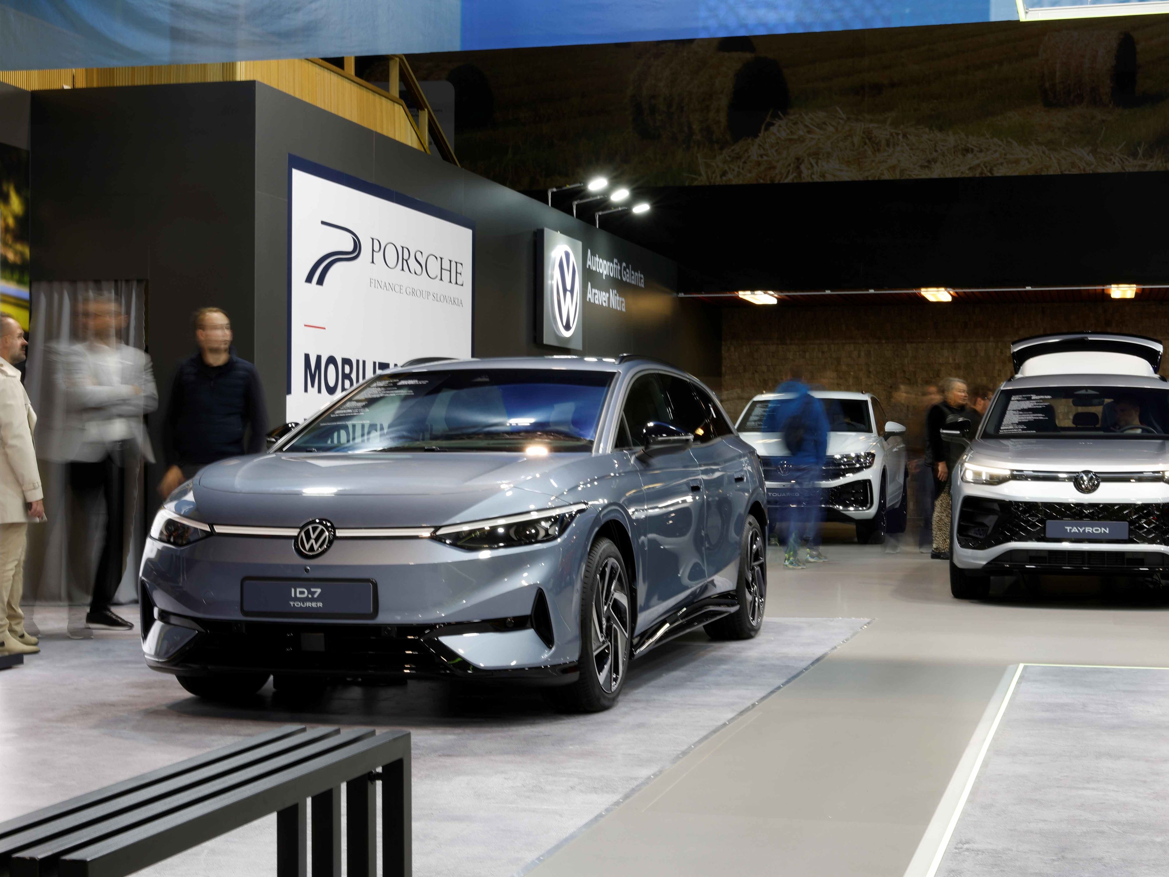 Na výstave sú prezentované elektrické vozidlá značiek Volkswagen. V popredí je modrý Volkswagen ID.7 a v pozadí biele SUV s otvoreným batožinovým priestorom. Okolo áut sa pohybujú návštevníci.