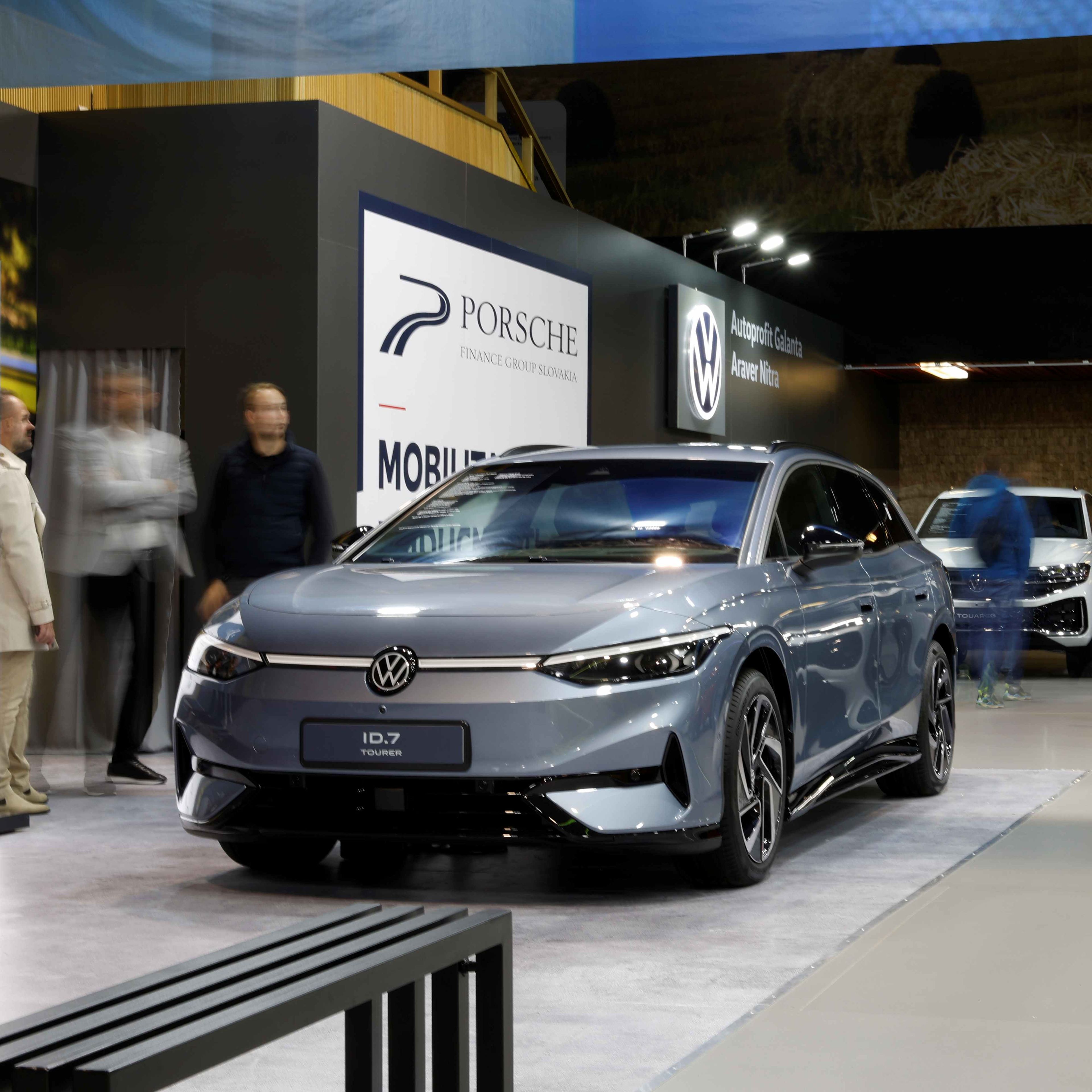 Na výstave sú prezentované elektrické vozidlá značiek Volkswagen. V popredí je modrý Volkswagen ID.7 a v pozadí biele SUV s otvoreným batožinovým priestorom. Okolo áut sa pohybujú návštevníci.