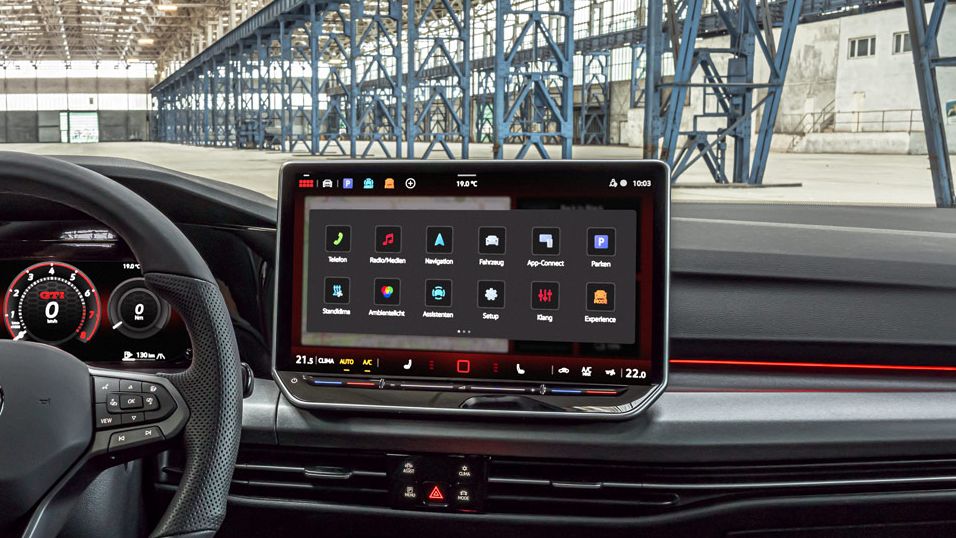Na obrázku je kokpit moderného modelu Golf GTI. Obzvlášť výrazný je veľký dotykový ovládací panel a digitálny prístrojový panel. V pozadí je vidieť veľkú priemyselnú budovu.