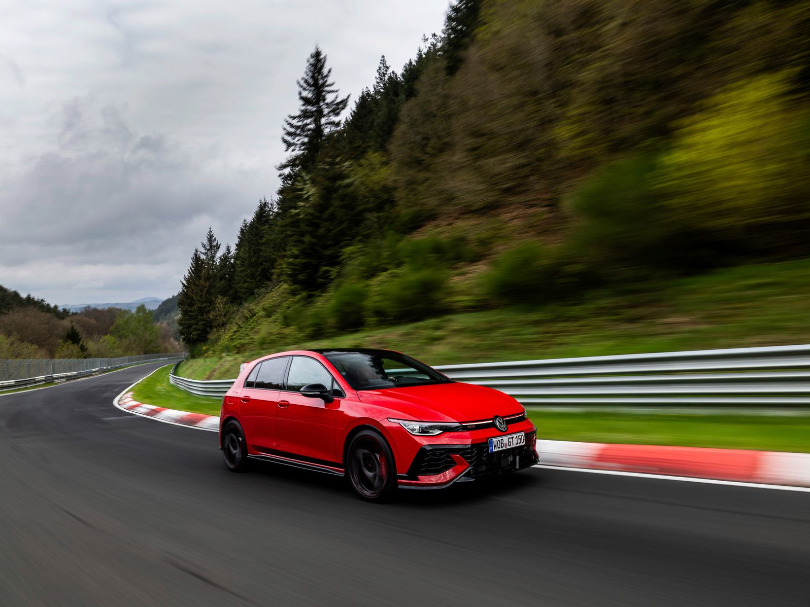 Červený Volkswagen Golf GTI prechádza zákrutou na závodnej trati obklopenej zeleňou a stromami. V pozadí sú viditeľné kopce a obloha je zamračená.