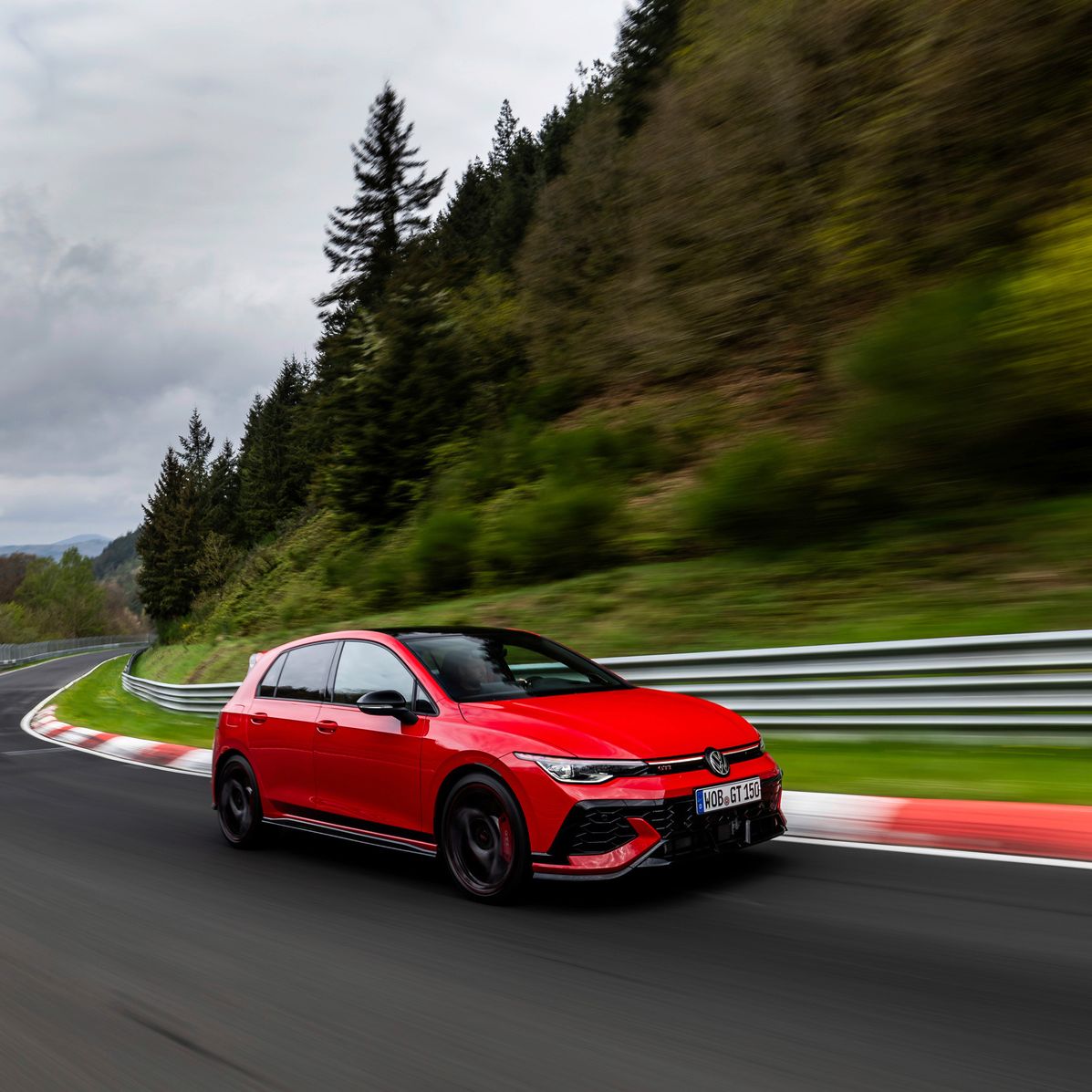 Červený Volkswagen Golf GTI prechádza zákrutou na závodnej trati obklopenej zeleňou a stromami. V pozadí sú viditeľné kopce a obloha je zamračená.