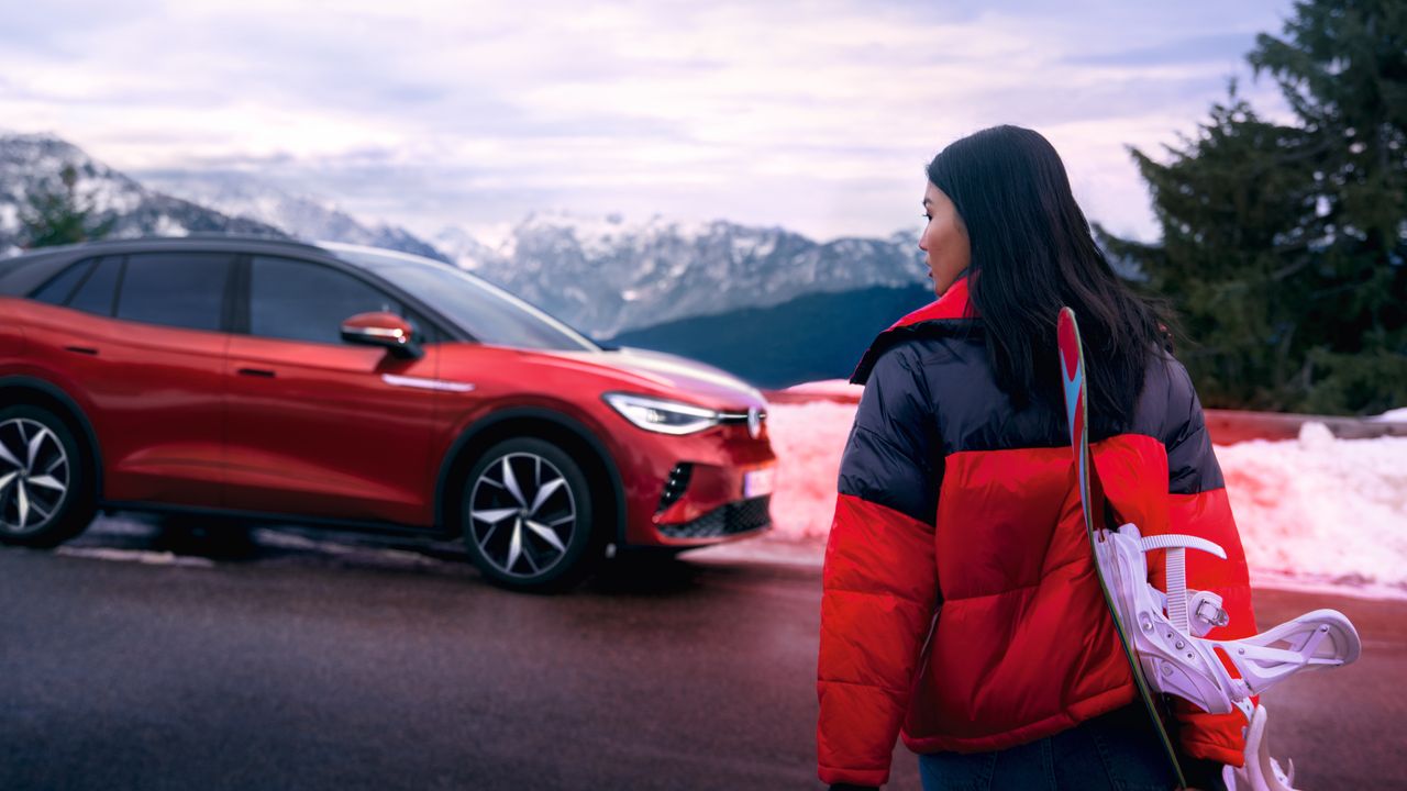 Bočný pohľad na červený VW ID.4 GTX na pozadí hôr, v popredí stojí žena, ktorá prichádza k vozidlu.