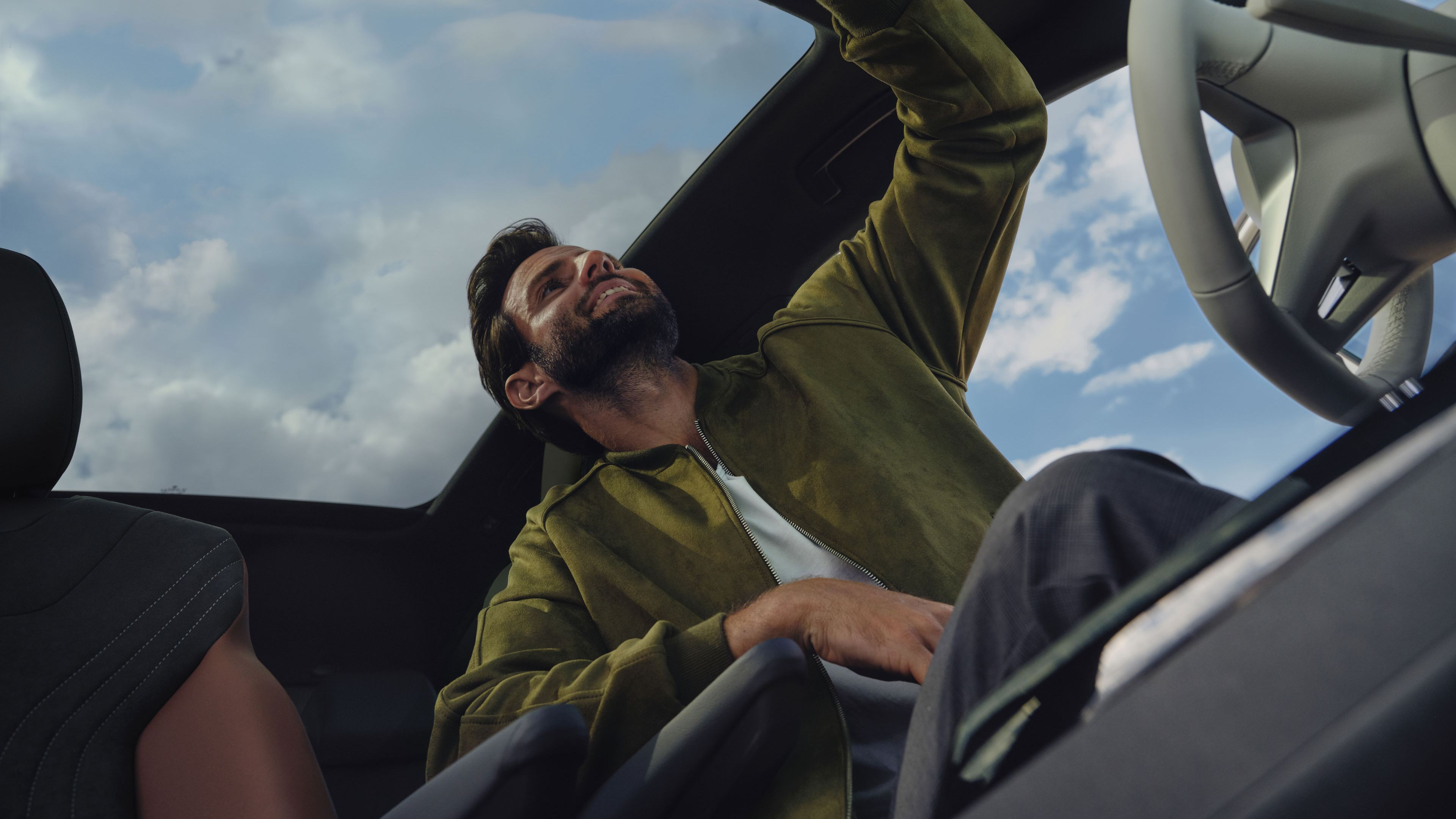 Muž v aute ukazuje prstom k ovládaciemu panelu v strope vozidla. Cez sklené panoramatické strešné okno vidieť mraky na oblohe.