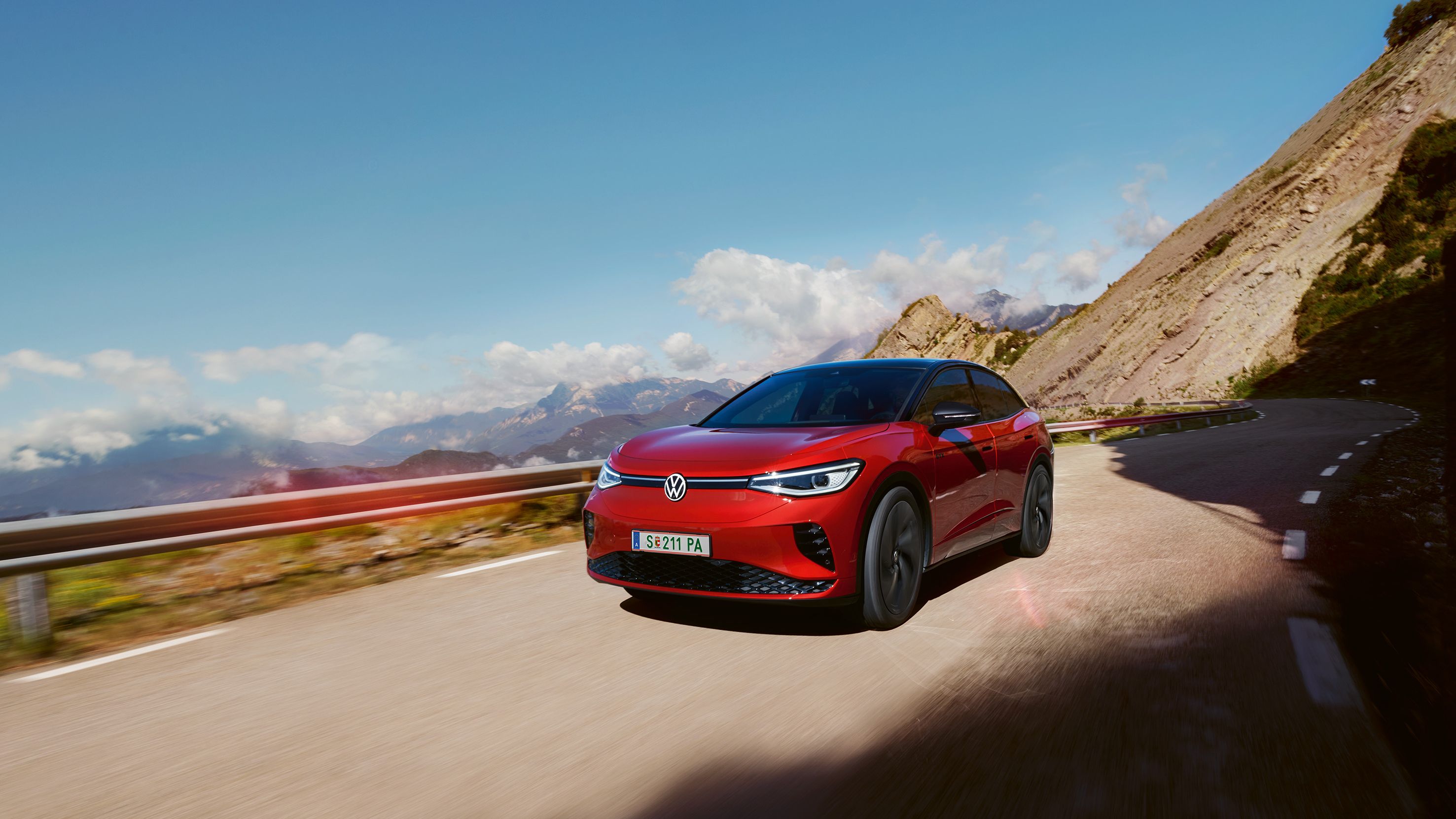 Červené Volkswagen ID.5 GTX  jazdí po ceste v horách. V pozadí vidieť ďalšie kopce zahalené do oblakov.