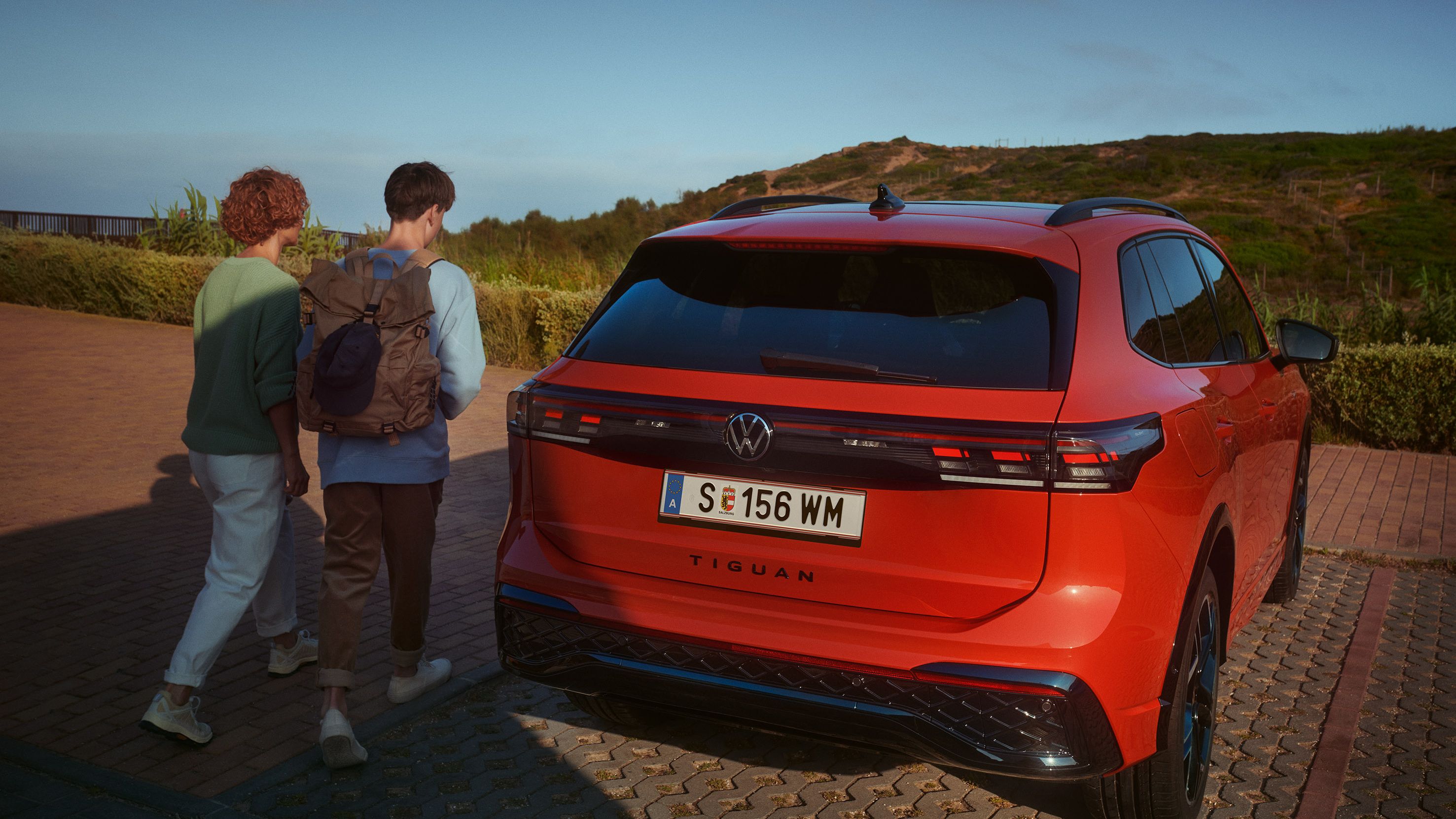 Pohľad na zadné partie zaparkovaného červeného VW Tiguan. Dvaja mladí ľudia sa približujú k vozidlu. V pozadí je kopec.