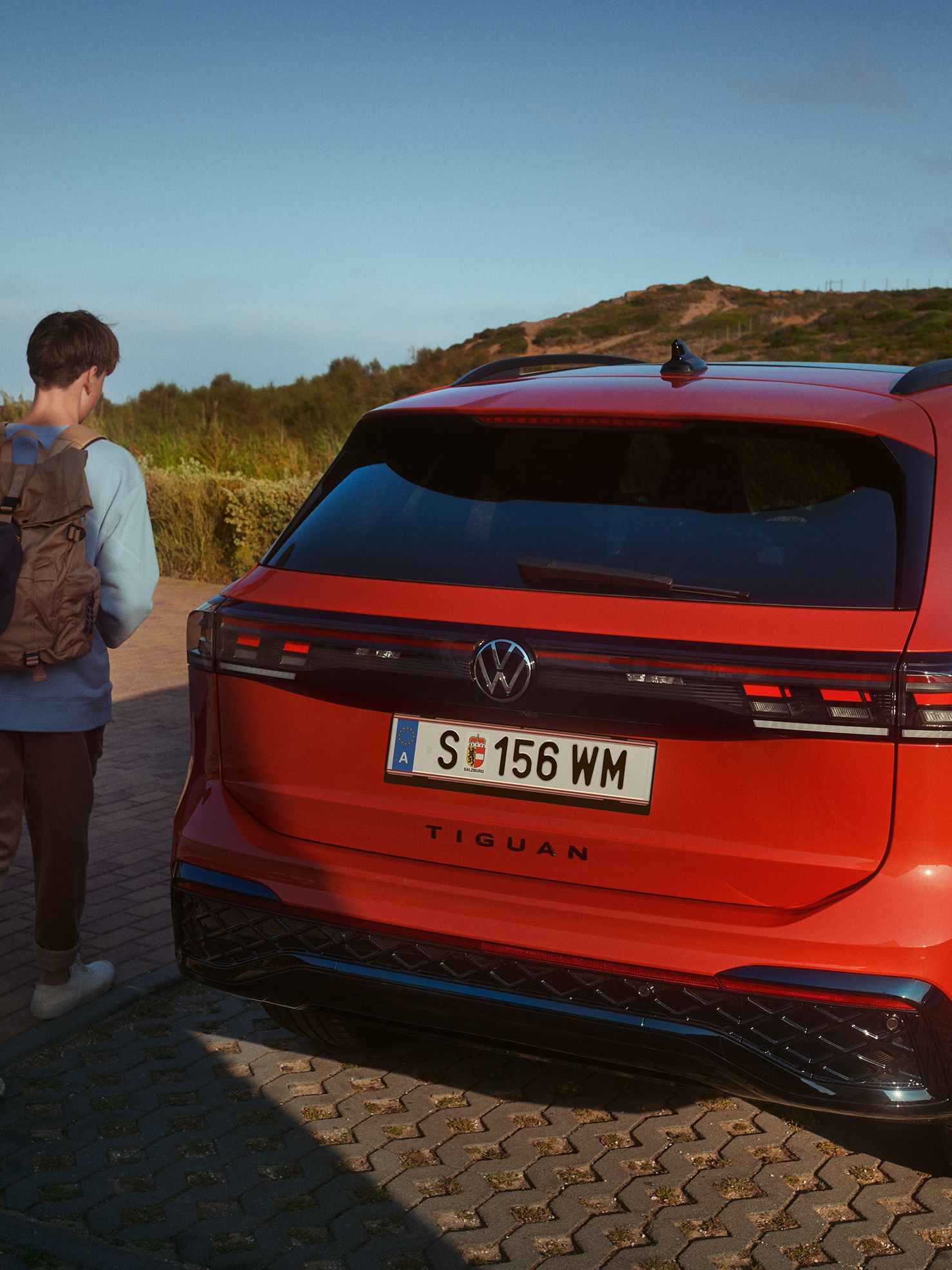 Pohľad na zadné partie zaparkovaného červeného VW Tiguan. Dvaja mladí ľudia sa približujú k vozidlu. V pozadí je kopec.