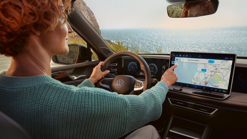 ,Žena s krátkymi, ryšavými kučeravými vlasmi sedí za volantom automobilu a používa dotykový displej s navigačnou mapou, na ktorej sú zobrazené trasy vedúce popri mori. Cez čelné sklo vidieť výhľad na oceán.