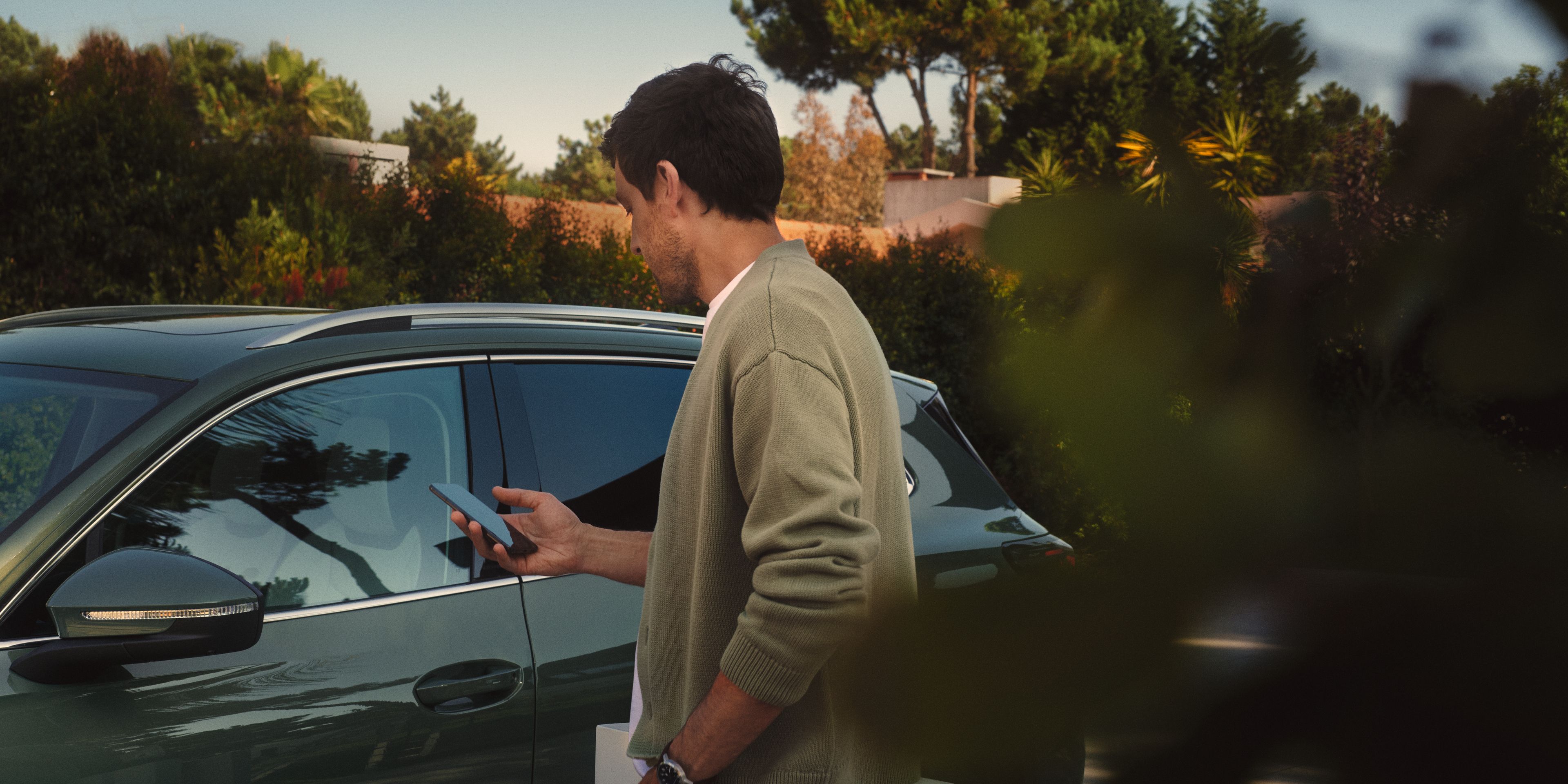 Muž stojí von pri zaparkovanom vozidle a pozerá sa na svoj mobil. V pozadí sú stromy a kríky. Je oblečený v svetlej bunde a rifliach.