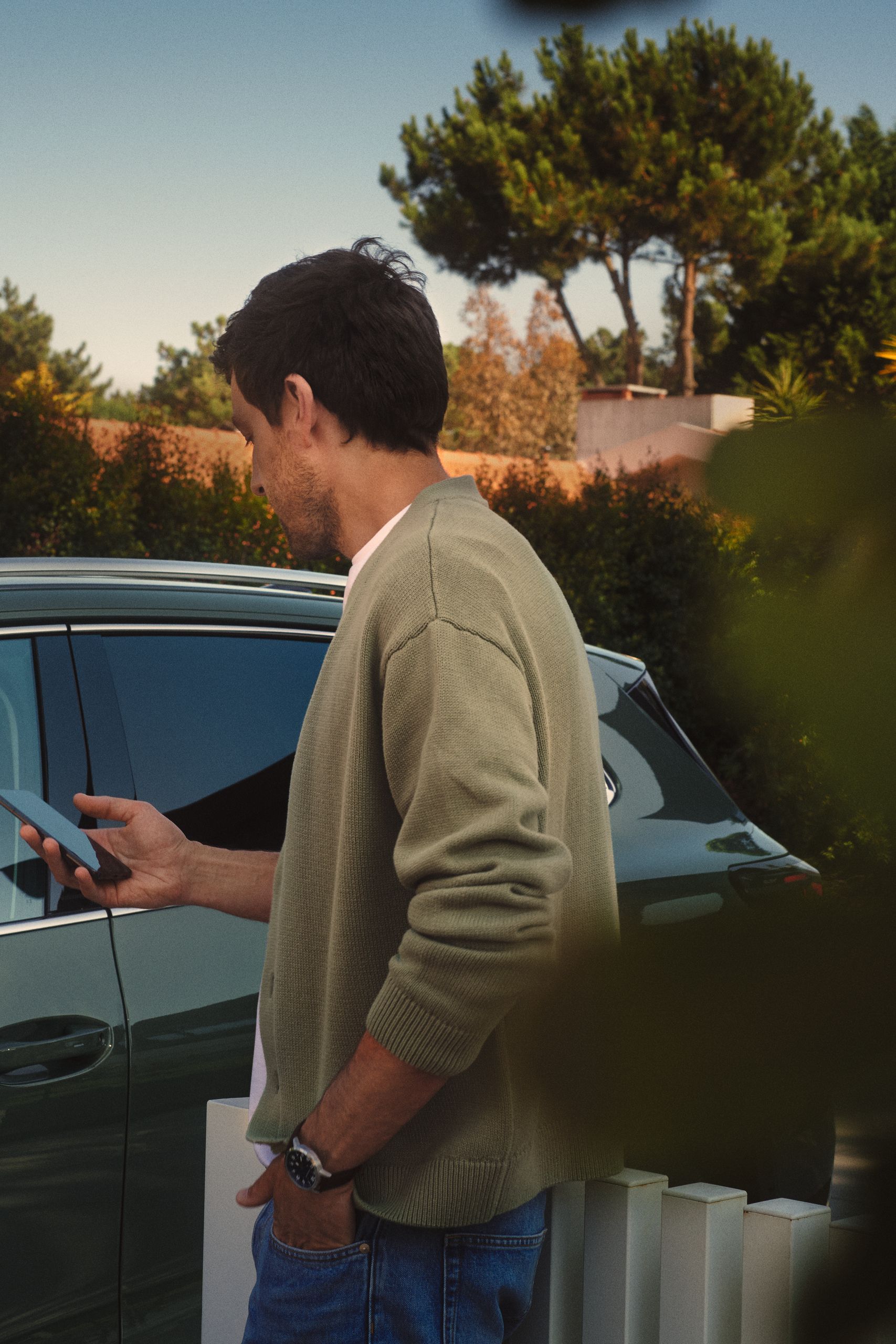 Muž stojí von pri zaparkovanom vozidle a pozerá sa na svoj mobil. V pozadí sú stromy a kríky. Je oblečený v svetlej bunde a rifliach.