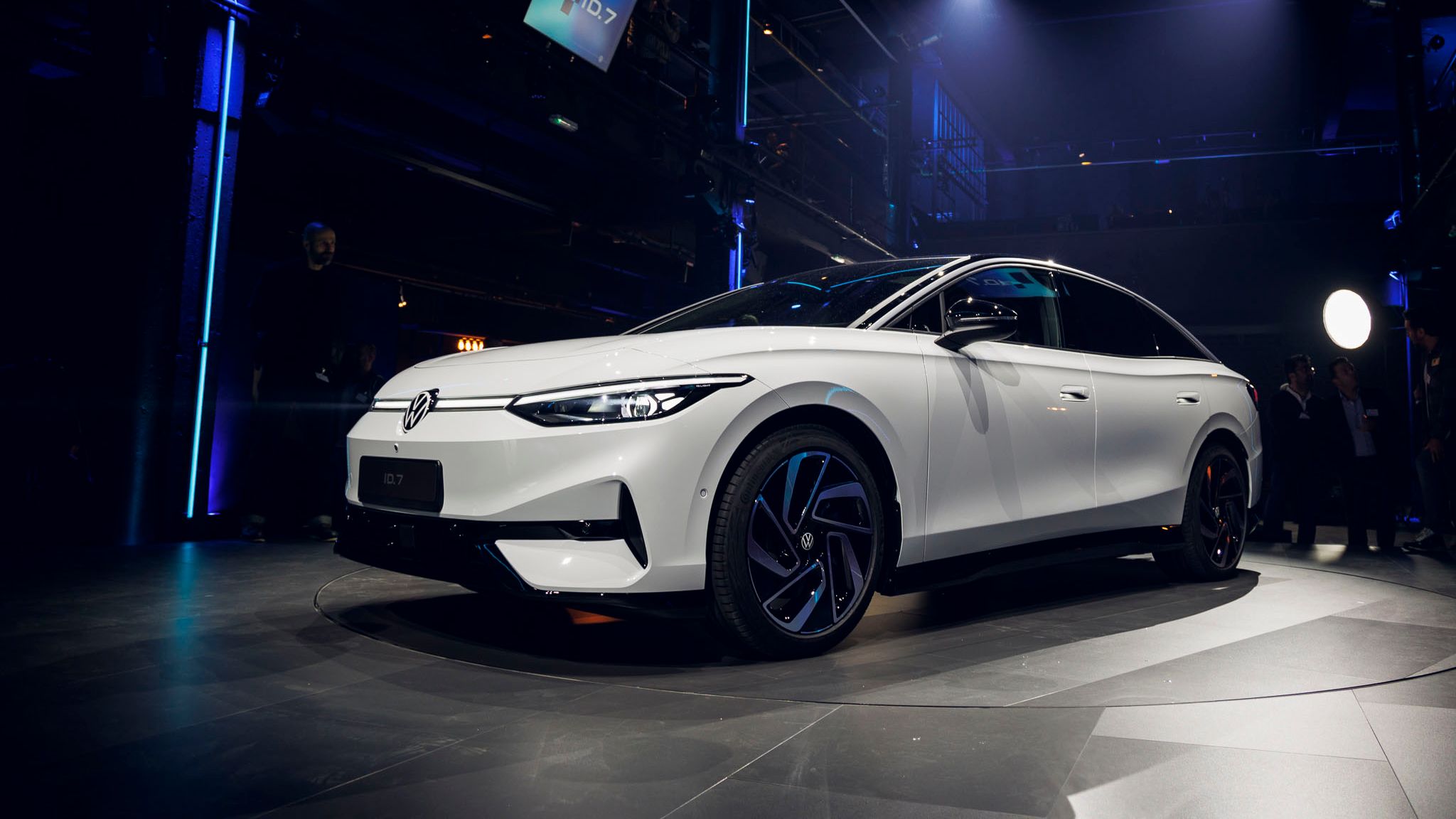 Moderný biely elektrický Volkswagen ID.7 stojí osvetlené na veľkom pódiu počas jeho uvedenia na trh v elegantnom prostredí. Naokolo sú modré svetlá a technické vybavenie. Na obrazovke v pozadí je viditeľné logo "ID.7".
