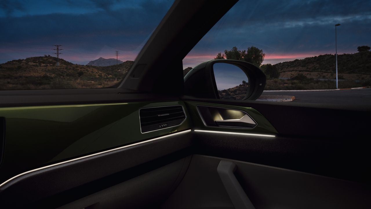 Pohľad do interiéru automobilu počas súmraku. Viditeľná časť palubnej dosky osvetlená ambientným osvetlením. Cez okná vidieť kopce, obloha je zatiahnutá a cez mraky presvitá ružový západ slnka.