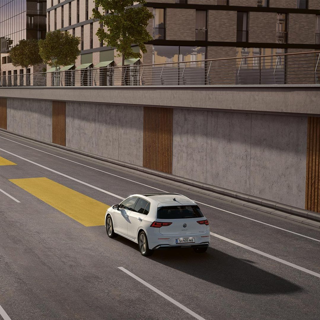 Biely VW Golf jazdí po ceste s aktívnym adaptívnym tempomatom ACC.  Vozidlo smeruje do tunela. V pozadí vidieť mesto pri zapadajúcom slnku.