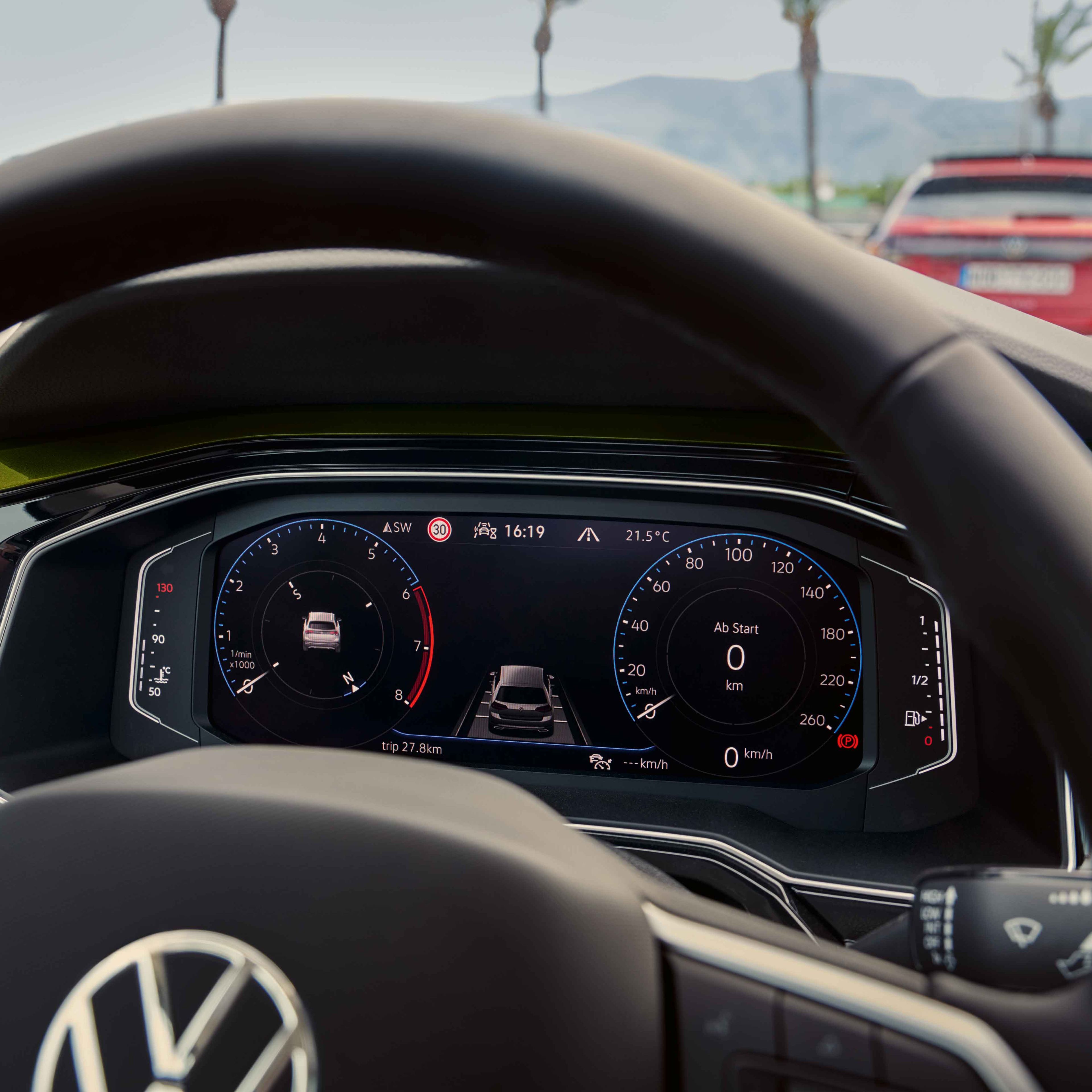 Digitálny prístrojový panel v modernom aute Volkswagen ukazuje informácie o jazde. V popredí vidieť volant s logom VW, cez čelné sklo vidieť pred autom rozmazané červené vozidlo. Na pozadí vidieť stromy a hory.