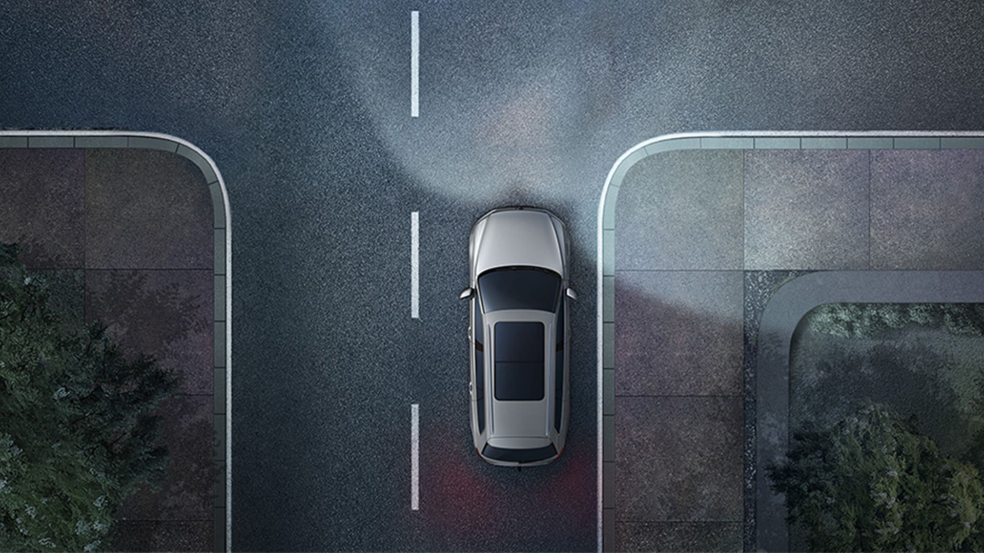 Automobil sa blíži ku križovatke v noci a  má zasvietené svetlá. Cesta je lemovaná stromami a chodníkmi. 