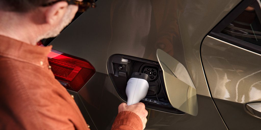 Muž v oranžovom saku nabíja elektrické auto. Nabíjací konektor drží v ruke a vkladá ho do nabíjacieho portu na aute. Auto je olivovo zelené s červeným zadným svetlom.