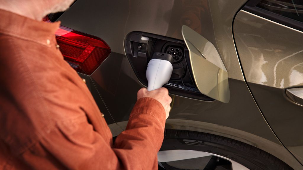 Muž v oranžovom saku nabíja elektrické auto. Nabíjací konektor drží v ruke a vkladá ho do nabíjacieho portu na aute. Auto je olivovo zelené s červeným zadným svetlom.