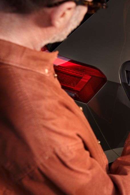 Muž v oranžovom saku nabíja elektrické auto. Nabíjací konektor drží v ruke a vkladá ho do nabíjacieho portu na aute. Auto je olivovo zelené s červeným zadným svetlom.