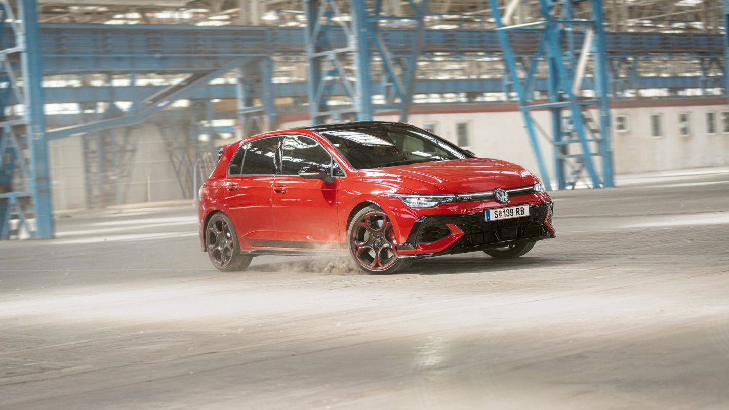 Červený Volkswagen Golf GTI sa preháňa prázdnou priemyselnou budovou. Pneumatiky víria prach. V pozadí sú viditeľné modré oceľové konštrukcie a okná.