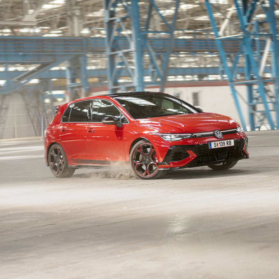 Červený Volkswagen Golf GTI sa preháňa prázdnou priemyselnou budovou. Pneumatiky víria prach. V pozadí sú viditeľné modré oceľové konštrukcie a okná.