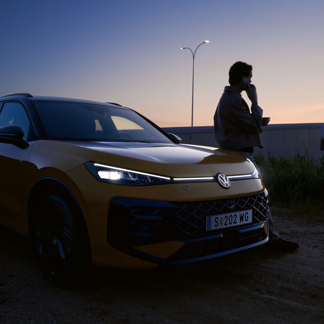 Muž sa opiera o vozidlo VW T-Roc R-Line so zapnutými svetlami a osvetleným logom.