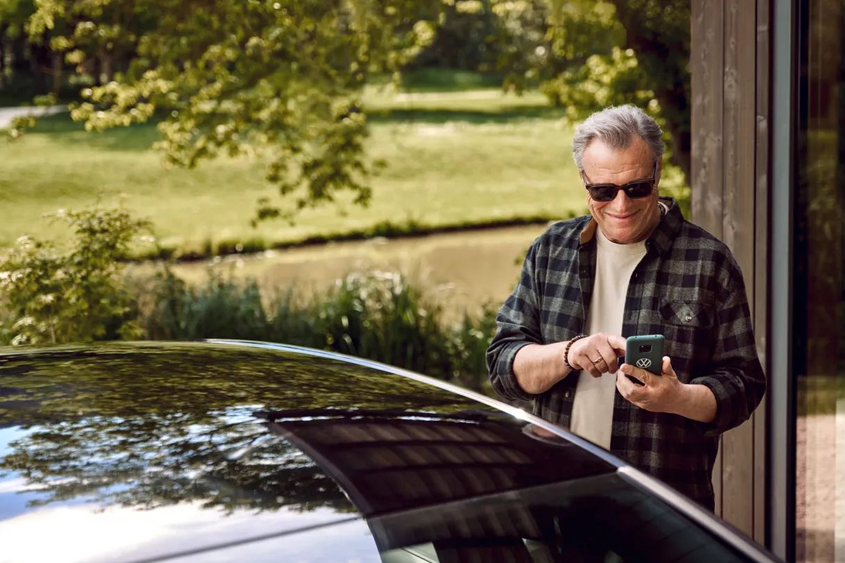 Muž stojí pri vozidle VW a drží v ruke mobilný telefón. Vozidlo je zaparkované pred domom, za ním je zelená lúka, v pozadí s chodníkom.