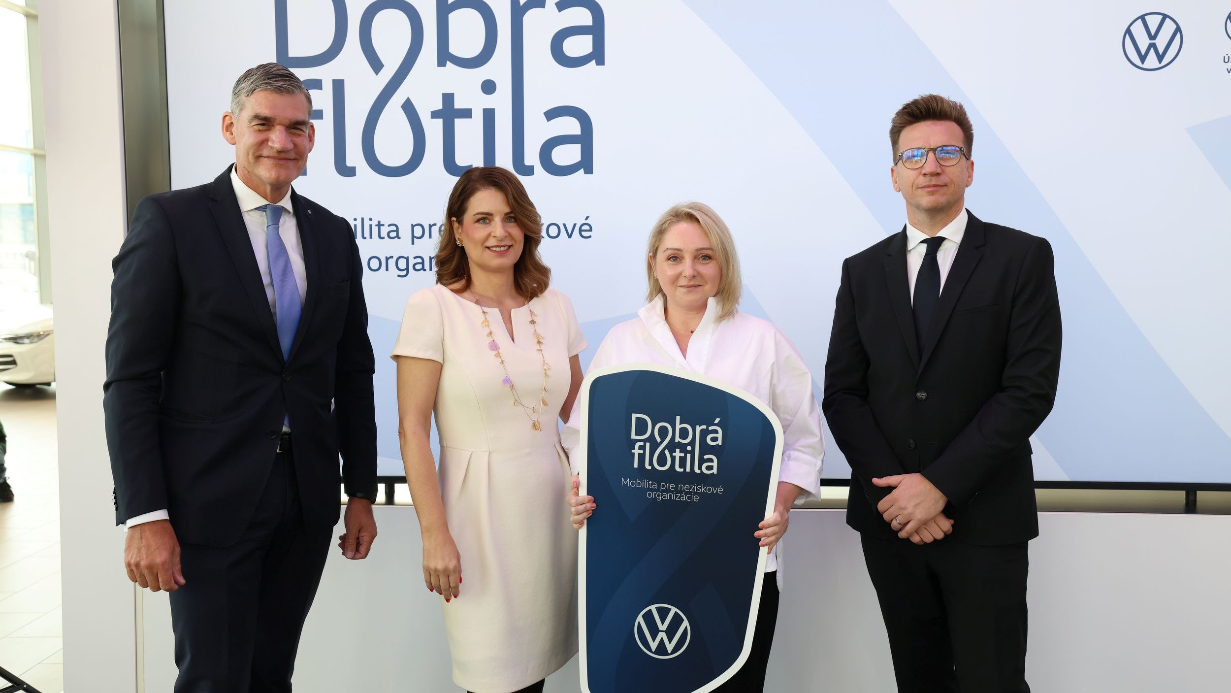 Štyria ľudia stojaci pred bannerom s logom "Dobrá flotila" a logom VW, pričom jeden z nich drží tabuľku s rovnakým logom.