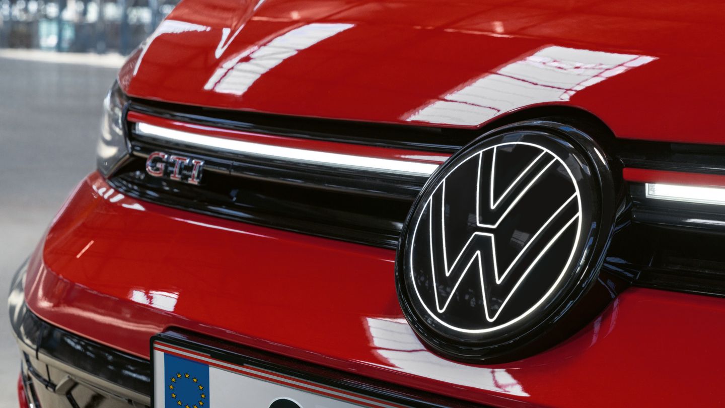 Detailný záber na prednú časť červeného modelu Golf GTI s nápisom GTI a charakteristickým logom VW. Auto je zaparkované v stredne veľkej garáži alebo hale.