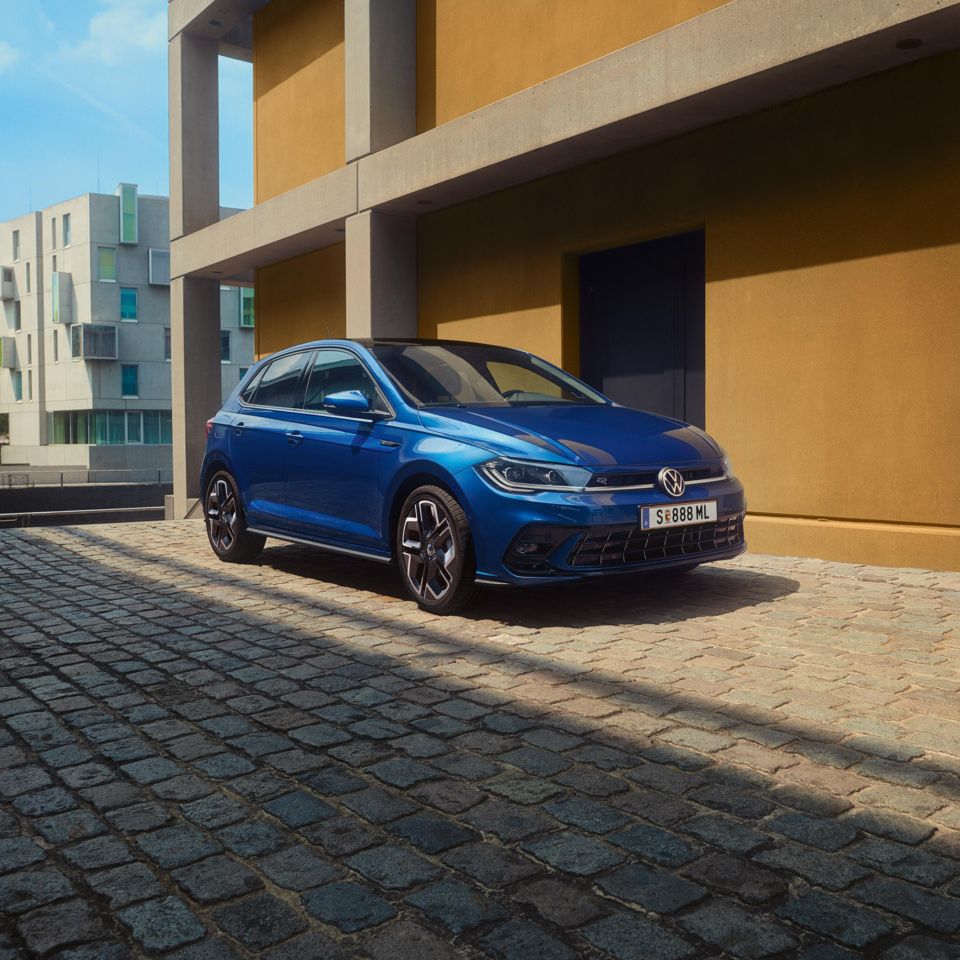 Modré Polo Volkswagen zaparkované na dláždenej ceste vedľa moderných bytových domov. V pozadí vidieť zeleň a zrejme cestu.