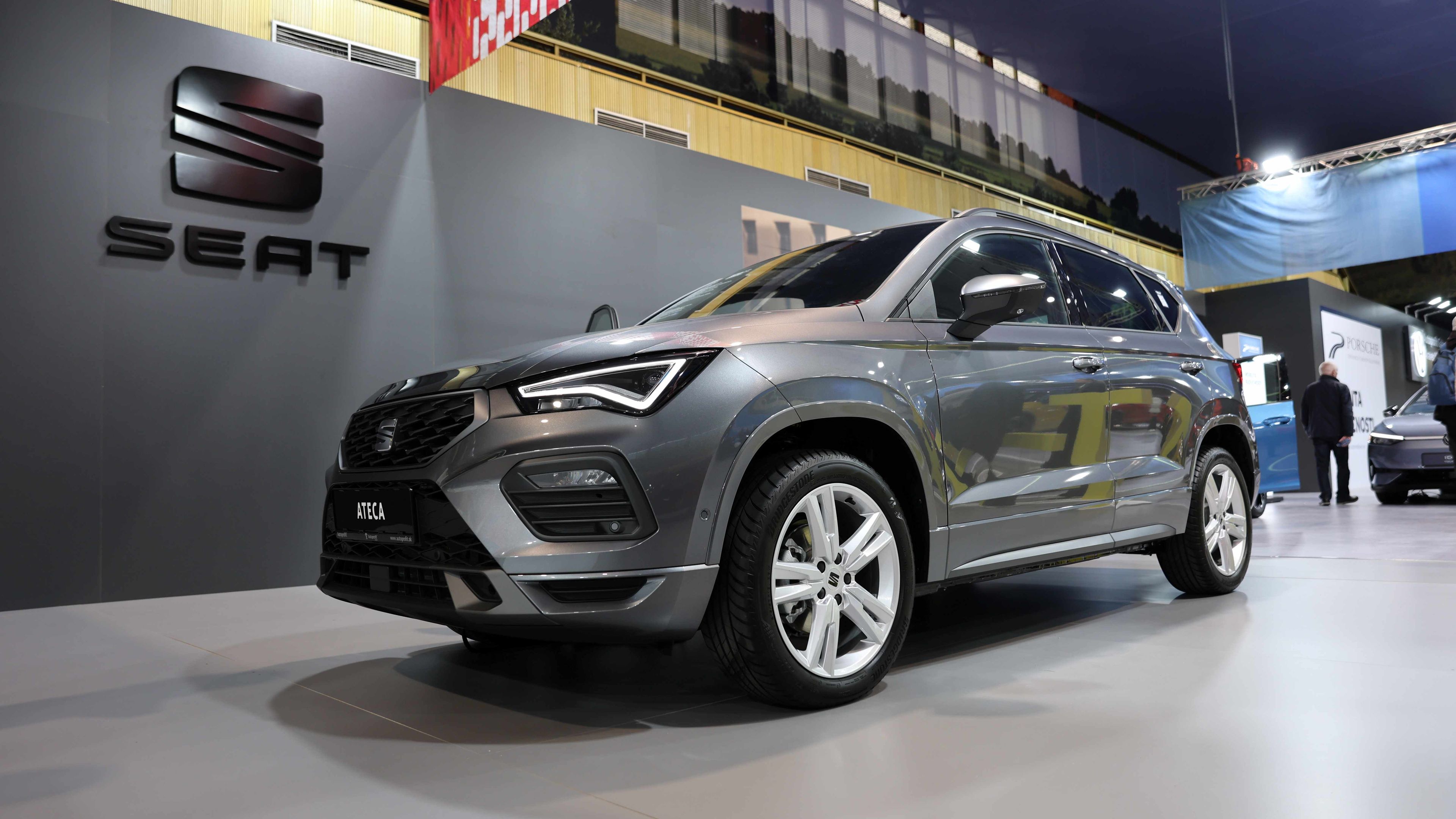 SEAT Ateca vystavené na autosalóne. V pozadí je vidieť stenu s logom SEAT a ďalší výstavný automobil. Scéna sa odohráva v modernom výstavnom priestore.