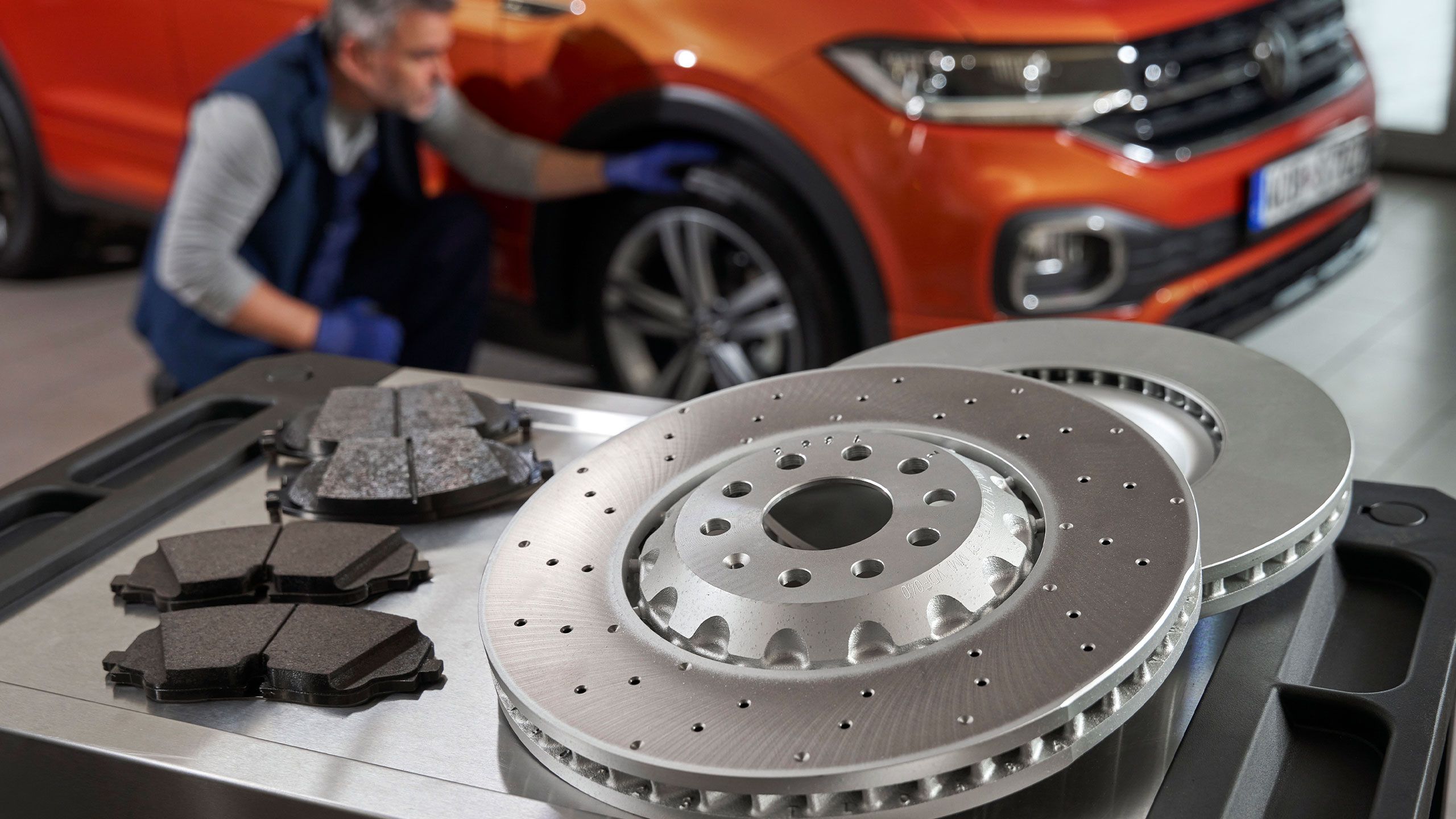 Mechanik kontroluje oranžové SUV, zatiaľ čo na stolíku sú umiestnené nové brzdové kotúče a brzdové doštičky.