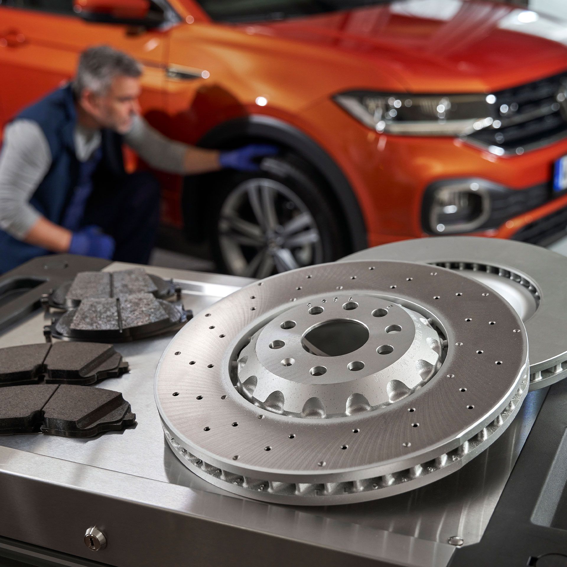 Mechanik kontroluje oranžové SUV, zatiaľ čo na stolíku sú umiestnené nové brzdové kotúče a brzdové doštičky.