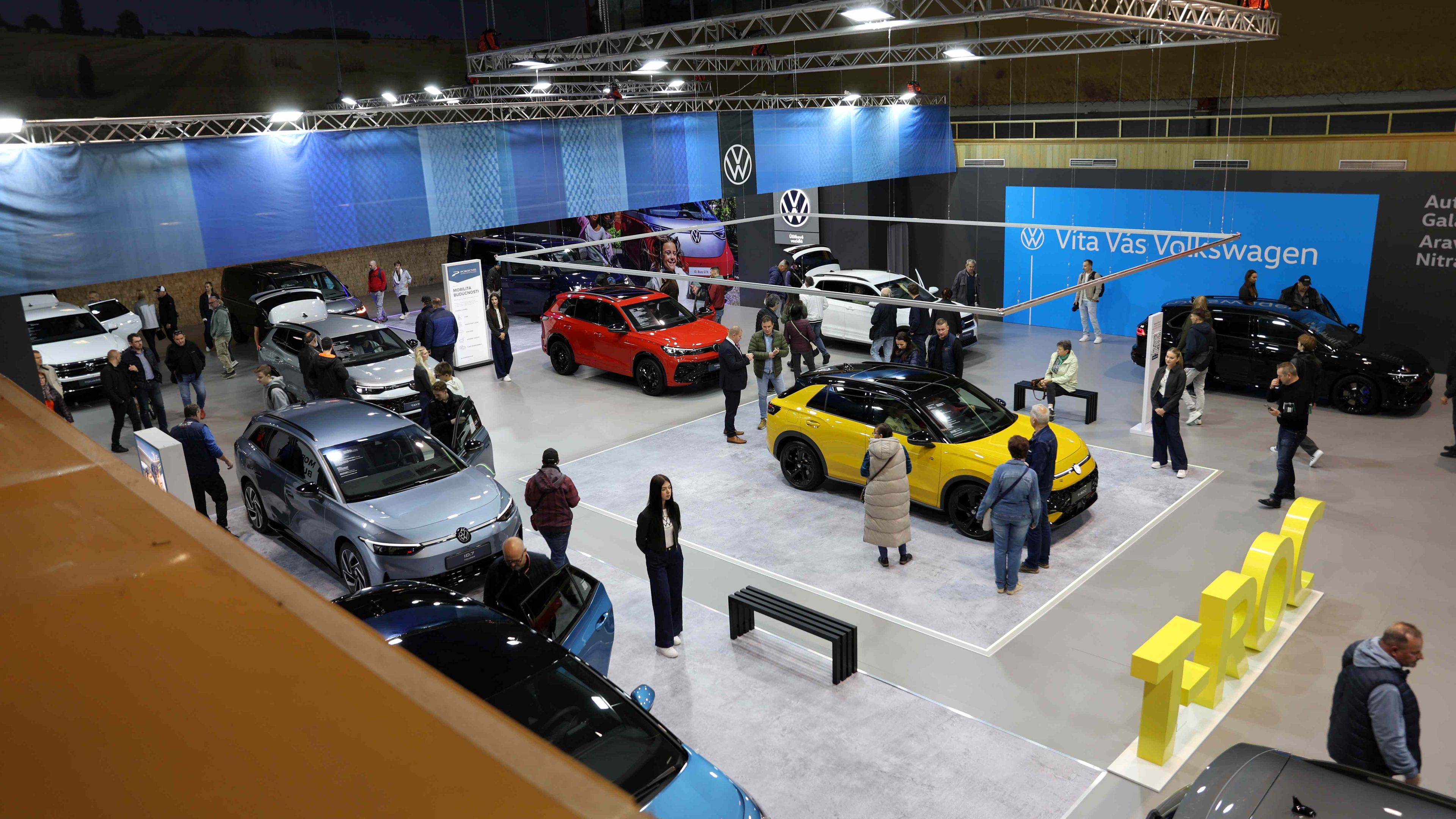 Výstavná hala plná ľudí obdivujúcich rôzne modely automobilov značky Volkswagen. Na popredí je jasne žlté auto. V pozadí je modrá stena s logom Volkswagen.