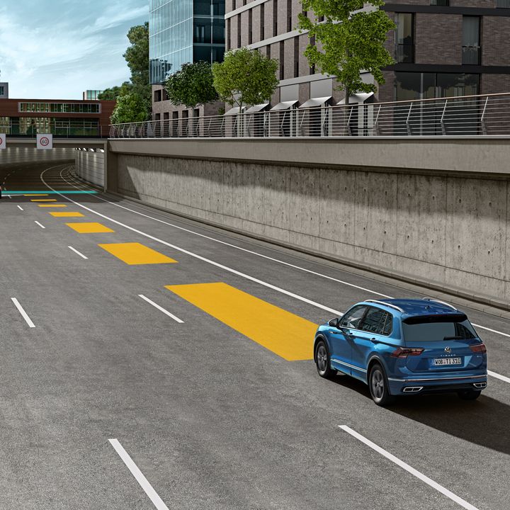 Automobil jazdí po ceste smerom do tunela v mestskej oblasti. Na ceste sú vyznačené žlté pruhy , ktoré znázorňujú adaptívny tempomat ACC. V pozadí sú moderné budovy a zeleň.