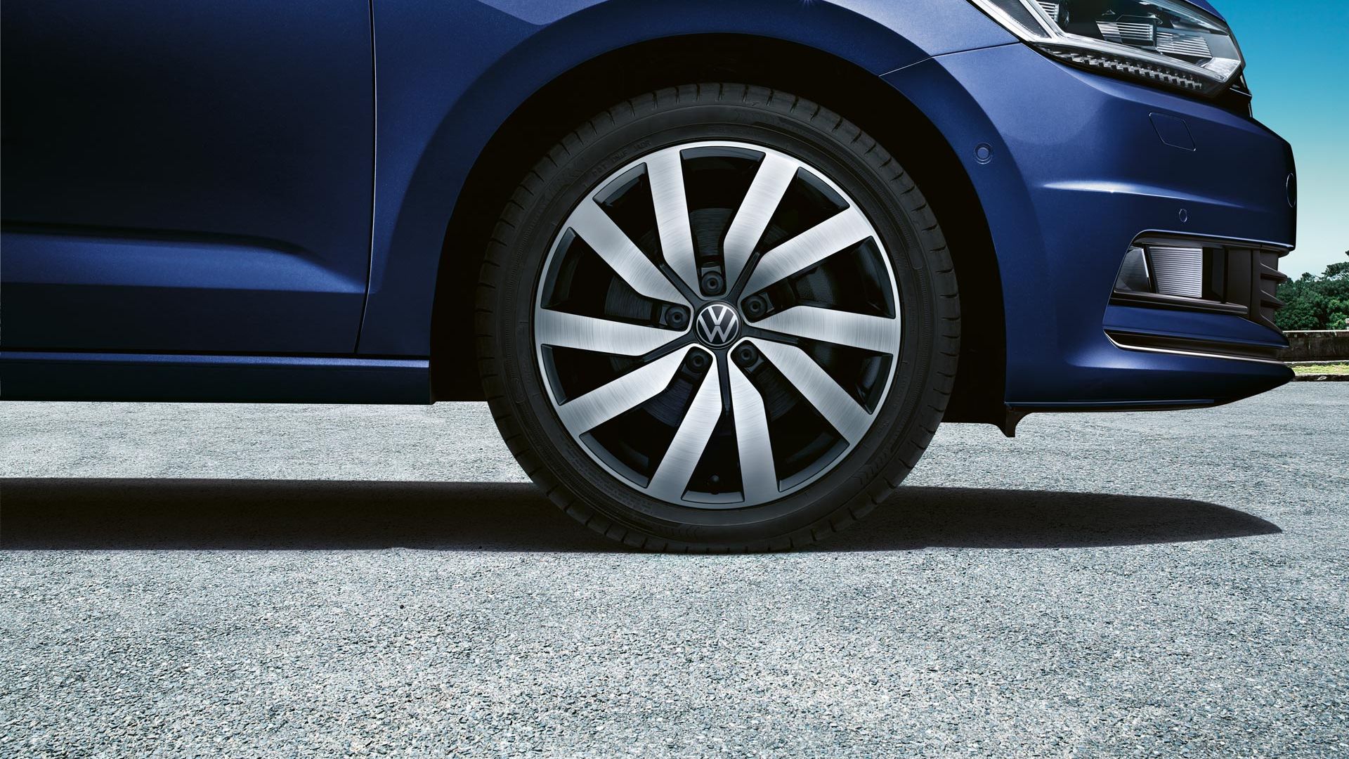 Detailný pohľad na prednú časť modrého vozidla Volkswagen, konkrétne na jeho koleso a disk. Vozidlo stojí na asfaltovej ceste. V pozadí je viditeľná časť krajiny s modrou oblohou.